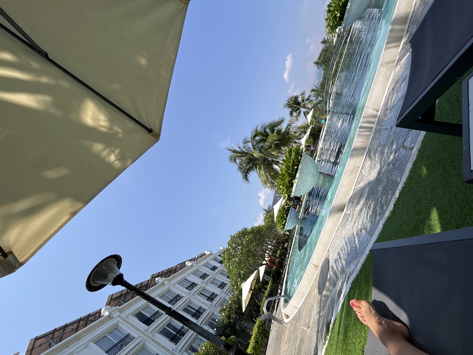 Here is a detailed description of the image:
The image captures a scene at a resort in Bac Nha Trang Ward, Vietnam, taken from a first-person perspective while lounging. A large, beige sun umbrella dominates the left side of the frame, casting shadows. Clear blue sky fills the background, punctuated by palm trees.
To the right is a long, multi-tiered swimming pool. The pool's edge is lined with light gray stone. Next to the pool is artificial green turf. A black sun lounger is positioned near the turf, and the person's leg is visible, with pink nail polish.
A lamppost extends from the bottom left, partially obscuring a multi-story white building. Green foliage is scattered among the buildings and the pool area.