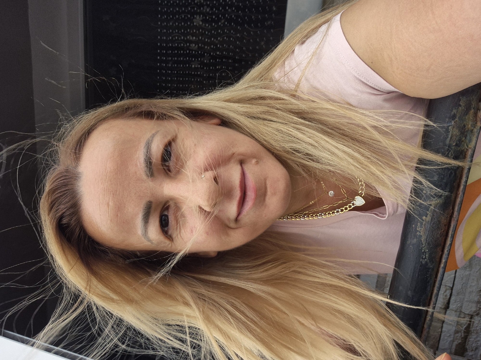 A medium shot, selfie-style photograph shows a woman with blonde hair and a light pink t-shirt. The woman is looking directly at the camera and smiling. Her hair is blown by the wind, obscuring parts of her face. She is wearing a gold necklace with a pendant shaped like a heart, and another pendant that appears to be a clear stone. A small mole is visible on her chin, and a nose ring is present on her left nostril. She is leaning against a dark, metallic railing, and behind her is a dark, patterned background. The overall lighting suggests an outdoor setting.