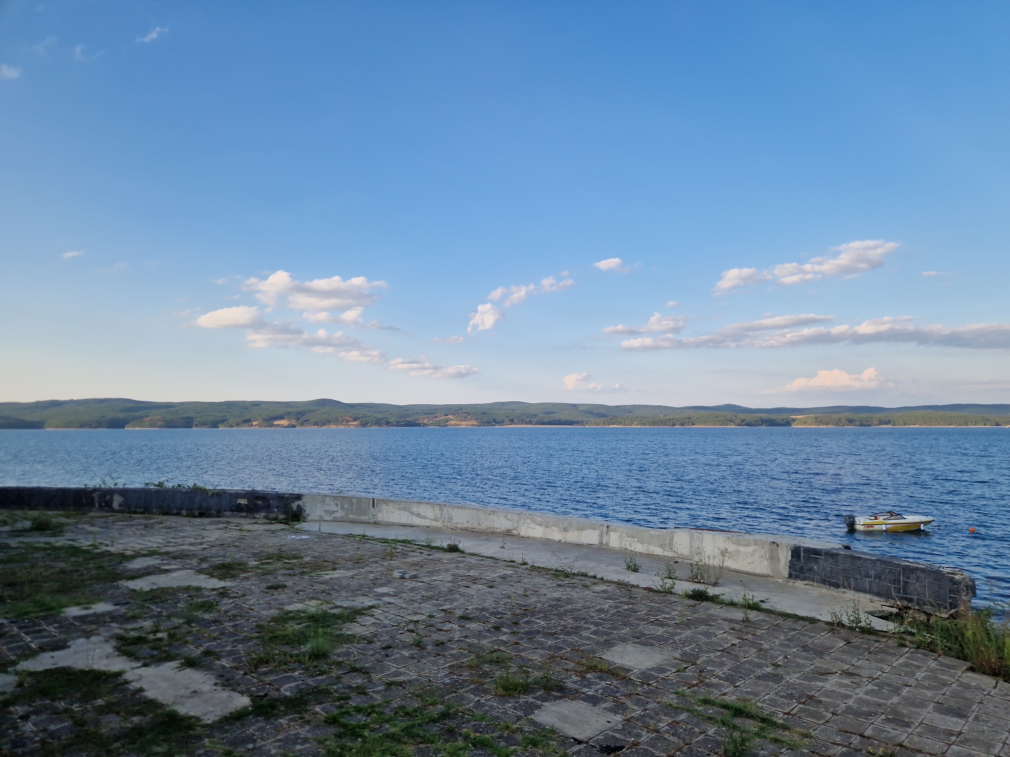 The image depicts a scenic view of the Pancharevo Lake, located near the village of Dolni Pasarel, Bulgaria. In the foreground, there is a paved area with interlocking square concrete tiles, some of which are overgrown with grass. A low, weathered concrete wall runs along the edge of this paved area, separating it from the lake. Beyond the wall, the calm blue water of the lake stretches towards a distant, forested shoreline. The sky above is a clear blue, dotted with scattered white clouds. A small, yellow and white motorboat is moored on the lake, slightly to the right of the frame. The overall atmosphere is tranquil and serene, suggesting a quiet day by the water.
