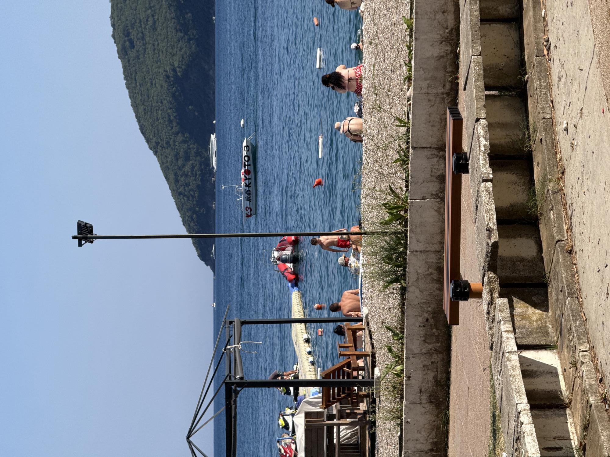 The image depicts a sunny day at a beach in Budva, Montenegro. In the foreground, there are concrete benches and a wooden bench. A stone retaining wall separates the seating area from a pebble beach. The beach is populated by several individuals, some sitting on the pebbles and others in the shallow water. A person is seated on a wooden chair under a shaded structure, with a table next to them. In the sea, there is a yellow floating dock with a red inflatable boat tied to it, and jet skis parked on the dock. Further out in the blue water, several boats are visible, including a white yacht and a smaller boat with the text "KYOTO 3" on its side. A green, tree-covered hill rises from the water in the background, characteristic of the Budva coastline. A lamppost stands near the center of the frame, positioned between the beach and the sea. The sky is clear and blue.