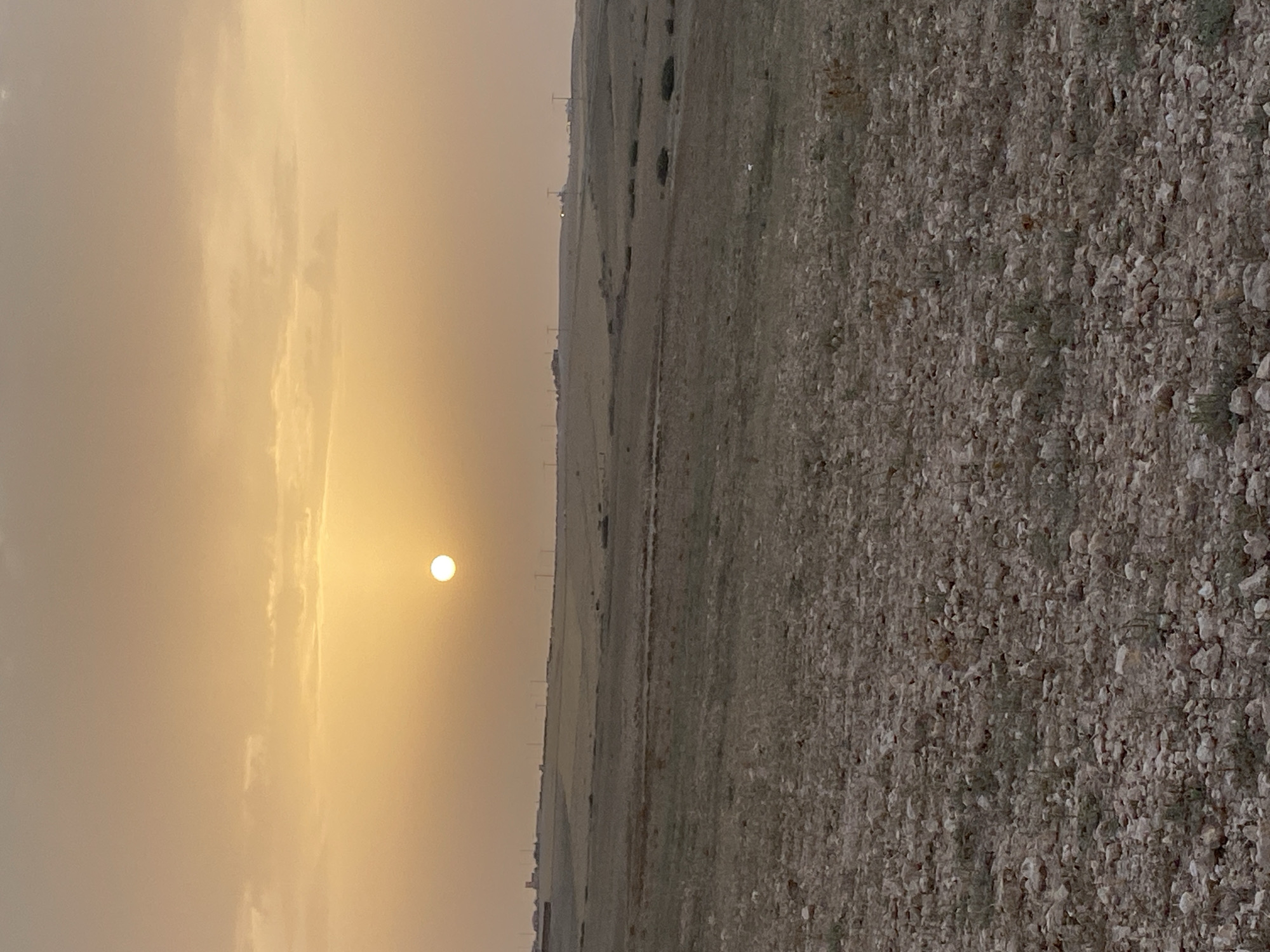 The image captures a landscape scene in Agafay, Morocco, during what appears to be sunset or sunrise. The sky is a soft, peachy-orange hue, with diffused sunlight visible as a bright, white orb near the center. Wispy clouds are scattered across the upper portion of the sky.
The terrain below is characterized by rolling hills covered in dry, sparse vegetation and scattered rocks. The foreground is dominated by a textured ground surface composed of numerous light-colored stones and pebbles, interspersed with dry grass and small plants. In the mid-ground, the landscape transitions to fields that appear more cultivated or naturally sparse, with patches of darker green vegetation visible. Further in the distance, along the horizon, a series of structures and utility poles are discernible, suggesting the presence of a settlement or infrastructure. The overall atmosphere is serene and evokes a sense of open, arid land.