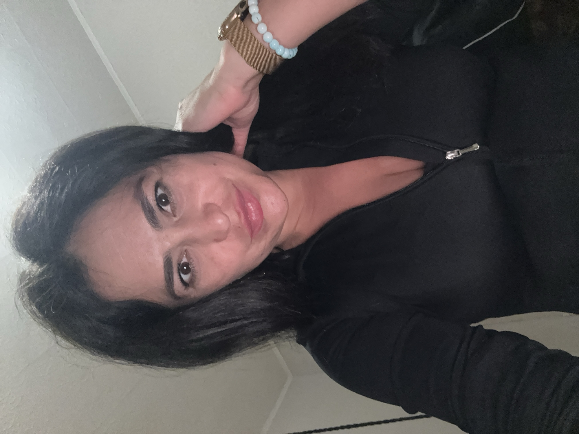 The image displays a close-up, rotated portrait of a woman. She has long, dark hair, dark eyes, and pronounced eyebrows. Her lips are a soft pink and curved into a gentle smile, suggesting a pleasant or content expression. She is wearing a dark, long-sleeved top, possibly black, with a silver zipper partially open near her chest.
On her right wrist, she has a gold-toned watch with a mesh band and a light blue beaded bracelet. Her right hand is raised, with her fingers touching the back of her head or running through her hair. The background is plain and light-colored, suggesting an indoor setting with artificial lighting. There are no distinct features in the background to indicate a specific location, though the context provided is Chaikovsky, Russia. No visible text is present in the image. The time of day and weather are not discernible from the indoor setting.