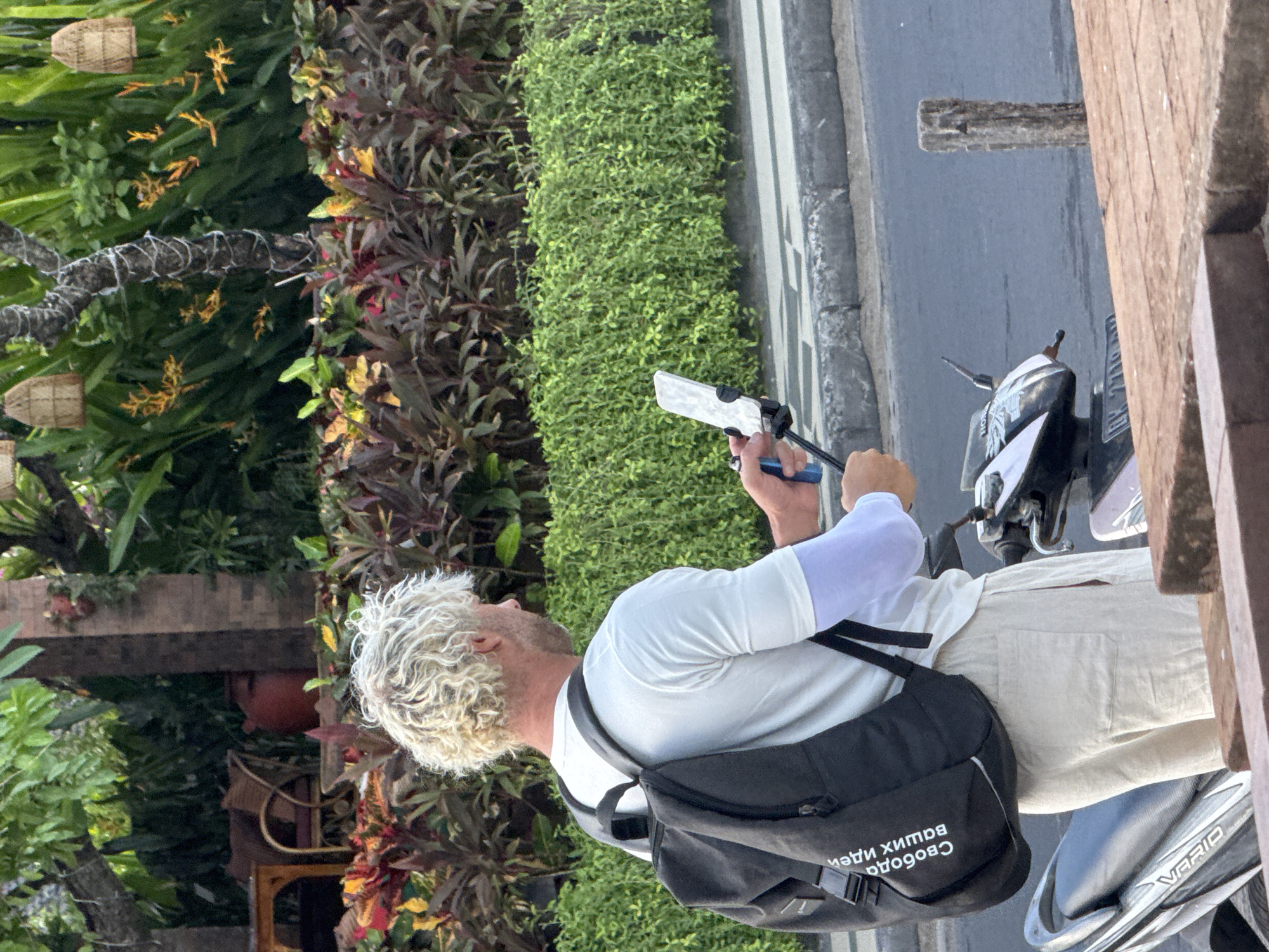 A blond-haired man wearing a white t-shirt, beige pants, and a black backpack is standing on a street in Kuta, Indonesia, holding a smartphone mounted on a selfie stick. He is wearing a light blue arm sleeve on his right arm. The man is facing away from the camera, looking to his right, and appears to be recording a video or taking a photo. In the background, there is lush green foliage, a row of colorful plants, and a tree decorated with string lights and bird nests. A scooter is parked to the man's right. The street has a curb and a paved surface. The image captures a sunny day with the sun casting shadows.