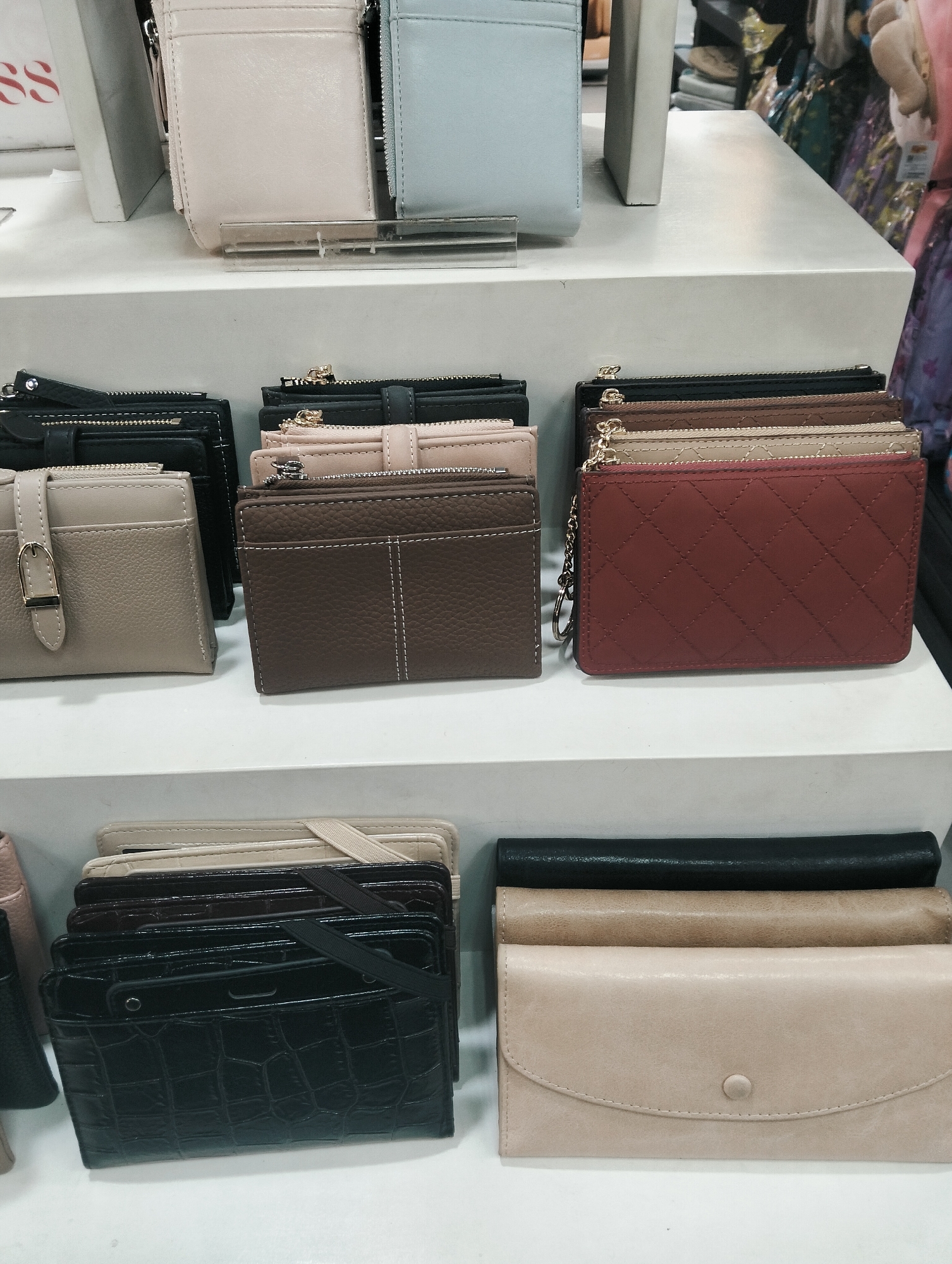 A retail display features a collection of wallets and coin purses neatly arranged on white shelves. On the top shelf, two wallets are visible: one is a light pink, and the other is a pale blue. The middle shelf showcases several coin purses and small wallets in various colors, including beige, brown, dark grey, and burgundy. The bottom shelf displays a stack of black, crocodile-patterned wallets and coin purses, along with several larger, flat wallets in beige and taupe colors. The environment appears to be a store, possibly in City of Medan, Indonesia, judging by the context. There are no people present in the image.