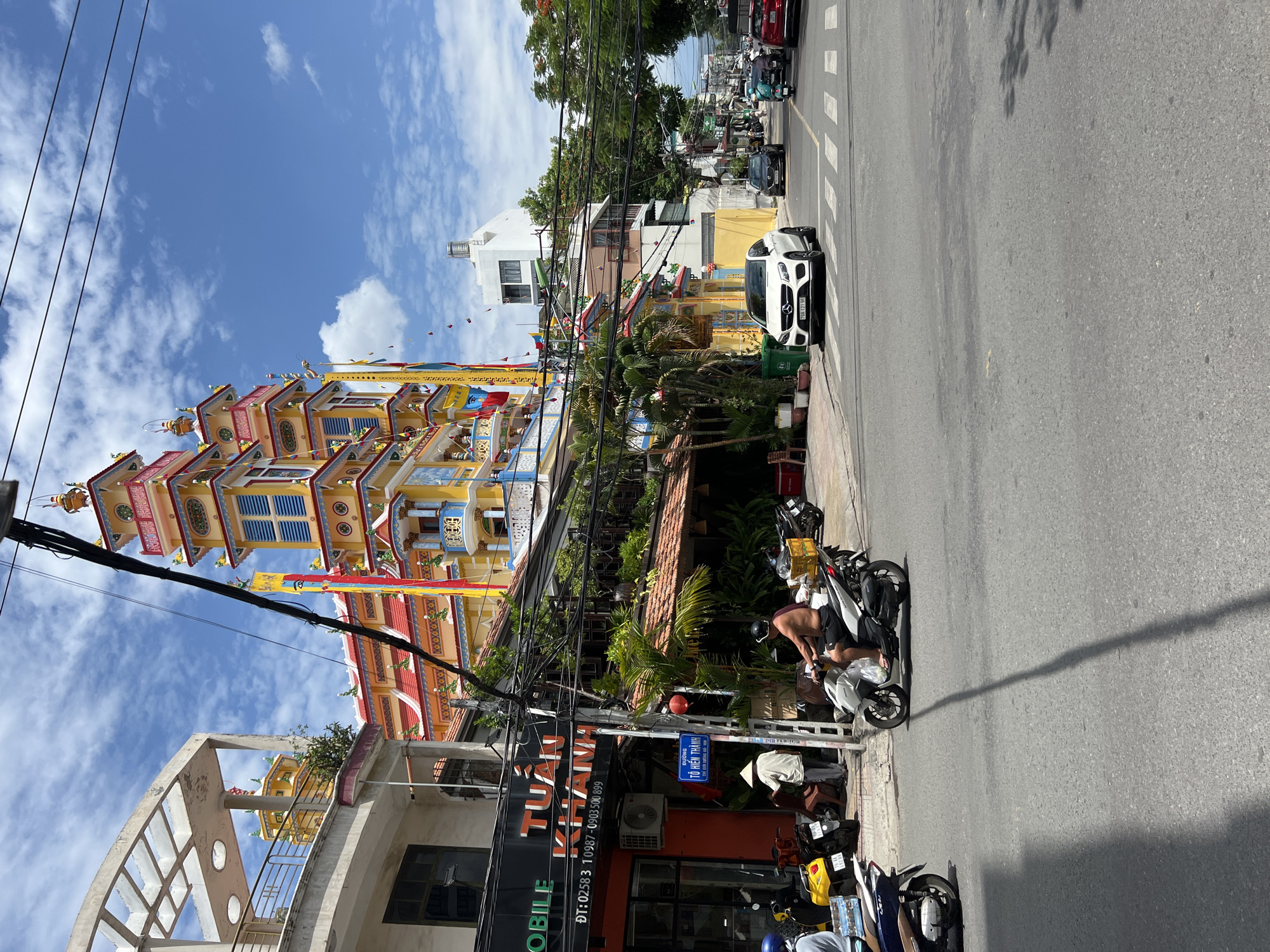 The image captures a street scene in Nha Trang, Vietnam, with a prominent colorful building in the background, possibly a temple or pagoda, adorned with ornate architectural details and flags. In the foreground, a man is riding a white scooter on the right side of the road. On the left side of the street, a yellow scooter and a dark blue scooter are parked. A person wearing a conical hat is standing near a building with signage that reads "TUẤN KHANH MOBILE". Several other vehicles, including cars and motorcycles, are visible on the road, and utility wires crisscross above. The sky is blue with scattered clouds, indicating a daytime scene. The architecture suggests a Southeast Asian urban environment, likely within Vietnam.