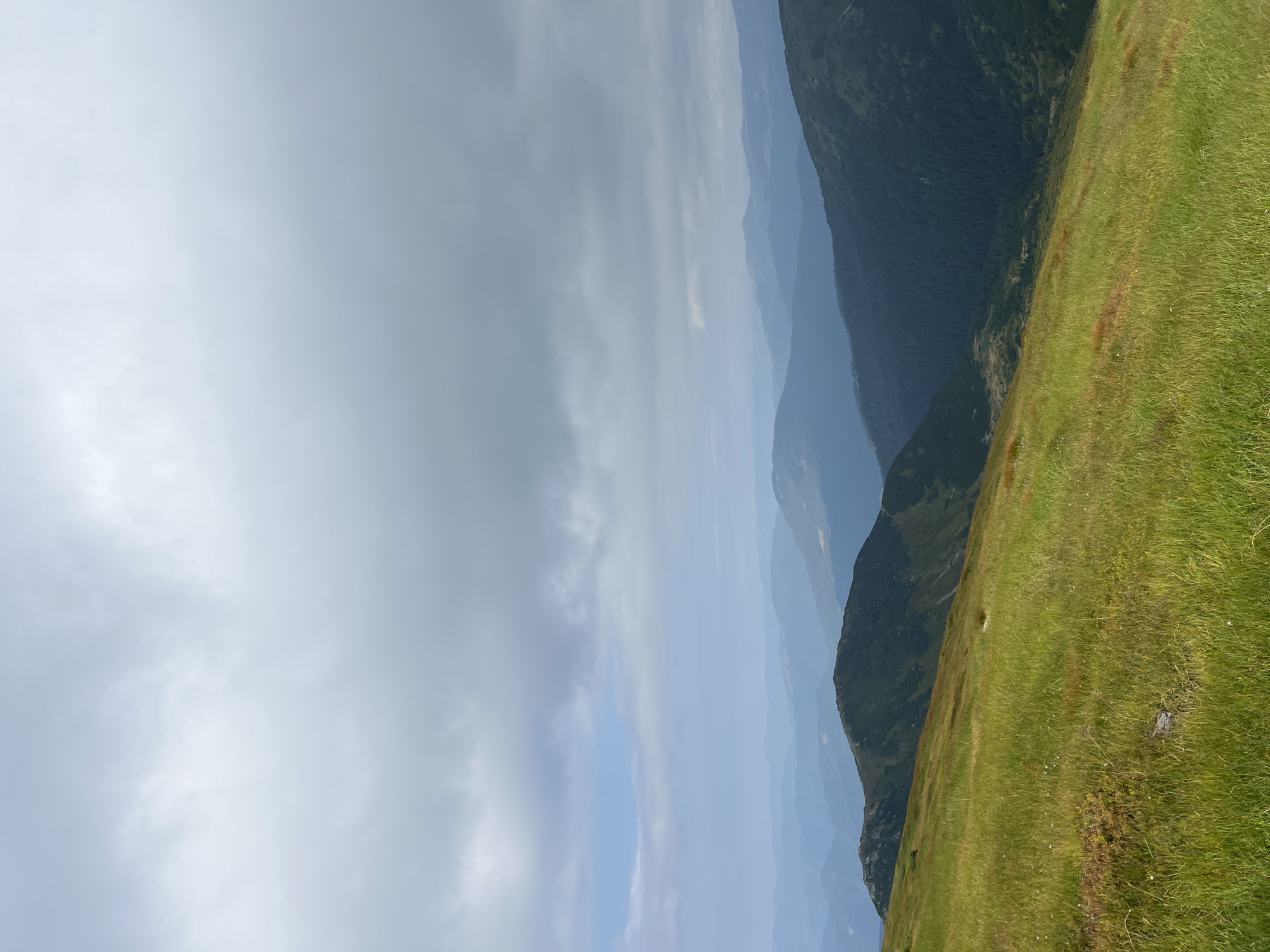 The image shows a panoramic view of rolling mountains in Luhy, Ukraine, under a cloudy sky. The foreground is dominated by a lush green meadow with tall grass, sloping downwards to the right. In the mid-ground, a dark green, forested mountainside descends into a valley. Beyond this, successive layers of hazy blue mountain ridges recede into the distance, creating a sense of depth. The sky is filled with heavy, grey clouds, with patches of lighter sky visible in the upper left quadrant. There are no people or man-made structures visible in the scene.