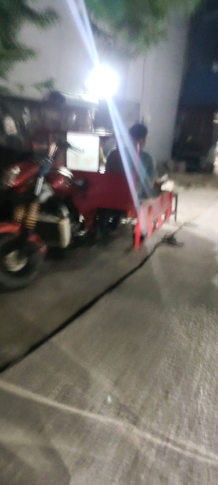 The image depicts a red three-wheeled vehicle, possibly a motorized rickshaw, parked on a paved surface that appears to be a street or walkway. The vehicle has a prominent front wheel and a covered rear section. A person, seen from the back, is seated in the driver's area, wearing a light-colored shirt. The lighting suggests it is nighttime or dusk, with bright light sources creating lens flares and illuminating the scene. Trees with foliage are visible in the background, and a building or wall is partially visible to the right. The overall image quality is low, with blurriness obscuring fine details. Based on the provided information, this scene is located in Mekarmukti, Indonesia.