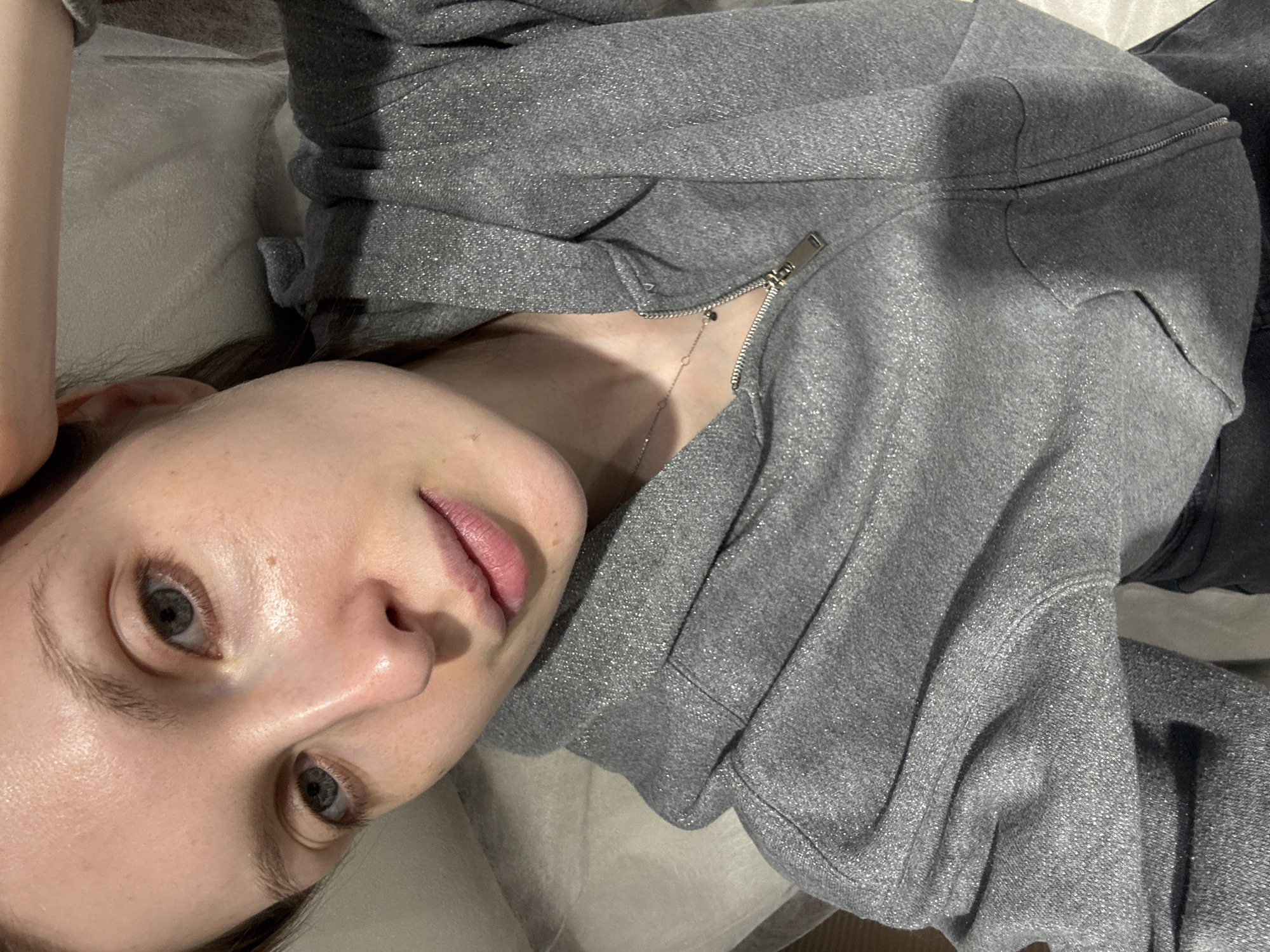 A person is captured in a close-up, upward-angled shot, lying down indoors. The image is rotated, presenting the subject's head turned towards the camera, with their body extending out of frame to the right. The individual has fair skin, light blue or grey eyes, and a natural, calm expression. Their lips are slightly parted, showing a natural pink tone. They appear to be wearing little to no makeup.
The person is dressed in a grey hooded top or jacket, distinguished by a shimmery or sparkly fabric that catches the light. The front zipper of the garment is partially undone, revealing a delicate, thin chain necklace with small, dark accents resting on their chest. Dark hair is subtly visible near the left ear, beneath the hoodie.
The background consists of a light-colored, soft-textured fabric, possibly a bedsheet or cushion, suggesting an indoor, relaxed setting. The lighting is soft and even, highlighting the details of the person's face and clothing. No specific activity is clearly depicted beyond posing for the photo. There is no visible text. The context indicates the location is Moscow, Russia.