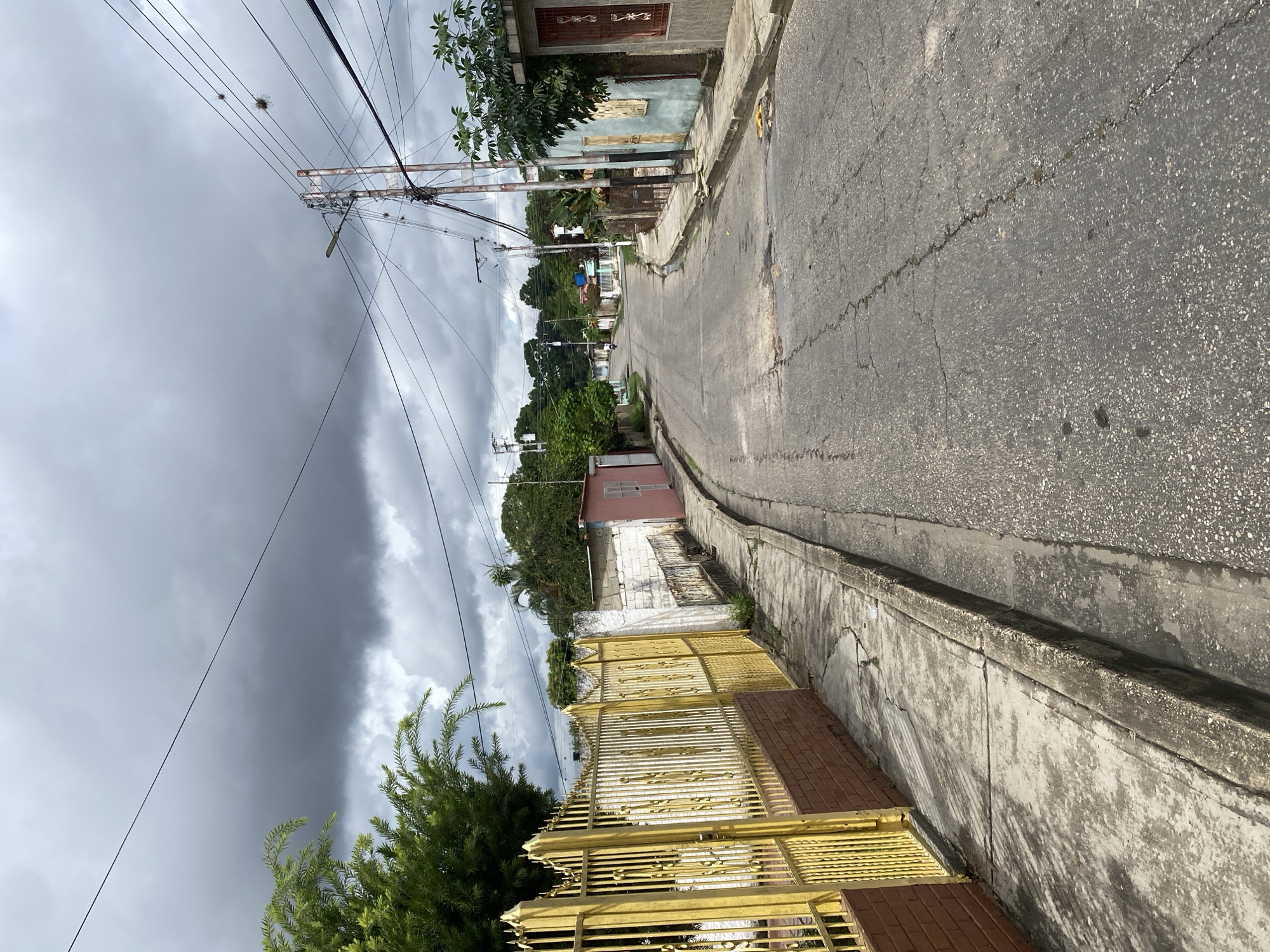 The image depicts a street scene in Tinaquillo, Venezuela. The view is down a slightly curving street, showing residential buildings on either side. The road surface is cracked and shows signs of wear. A concrete curb runs along the right side of the road. Power lines are visible overhead, crisscrossing the sky. On the left, a gold-colored metal fence stands in front of a building with a reddish-brown brick base. The sky is overcast with dark, ominous clouds. Vegetation includes trees and bushes along the street and near the buildings. No people or animals are immediately visible.