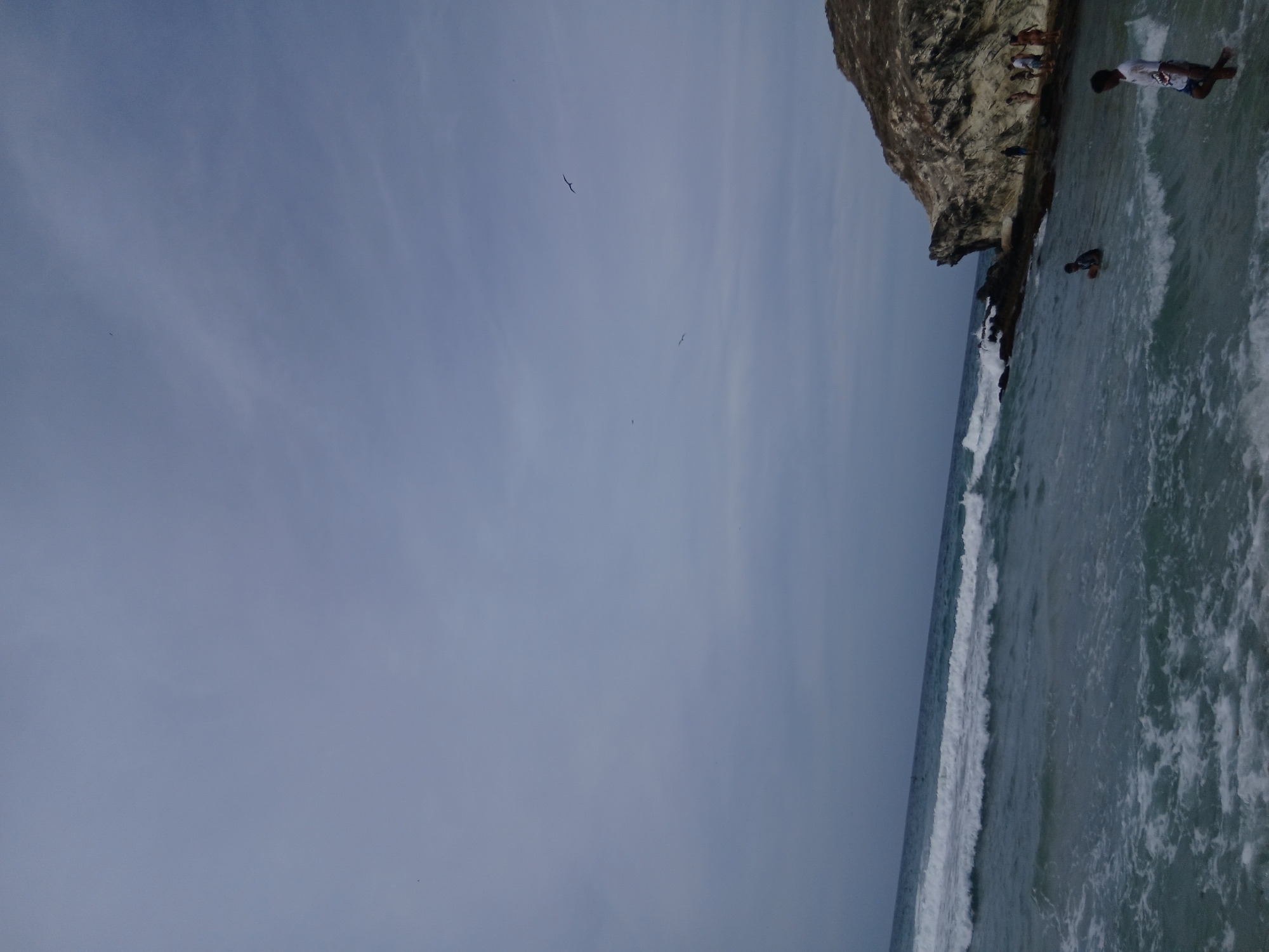 The image shows a beach scene at Playa Parguito, Venezuela. A rocky cliff with several people standing on it is visible on the right. Two individuals are partially submerged in the water. Waves are breaking along the shoreline. The sky is overcast with a few birds flying overhead.