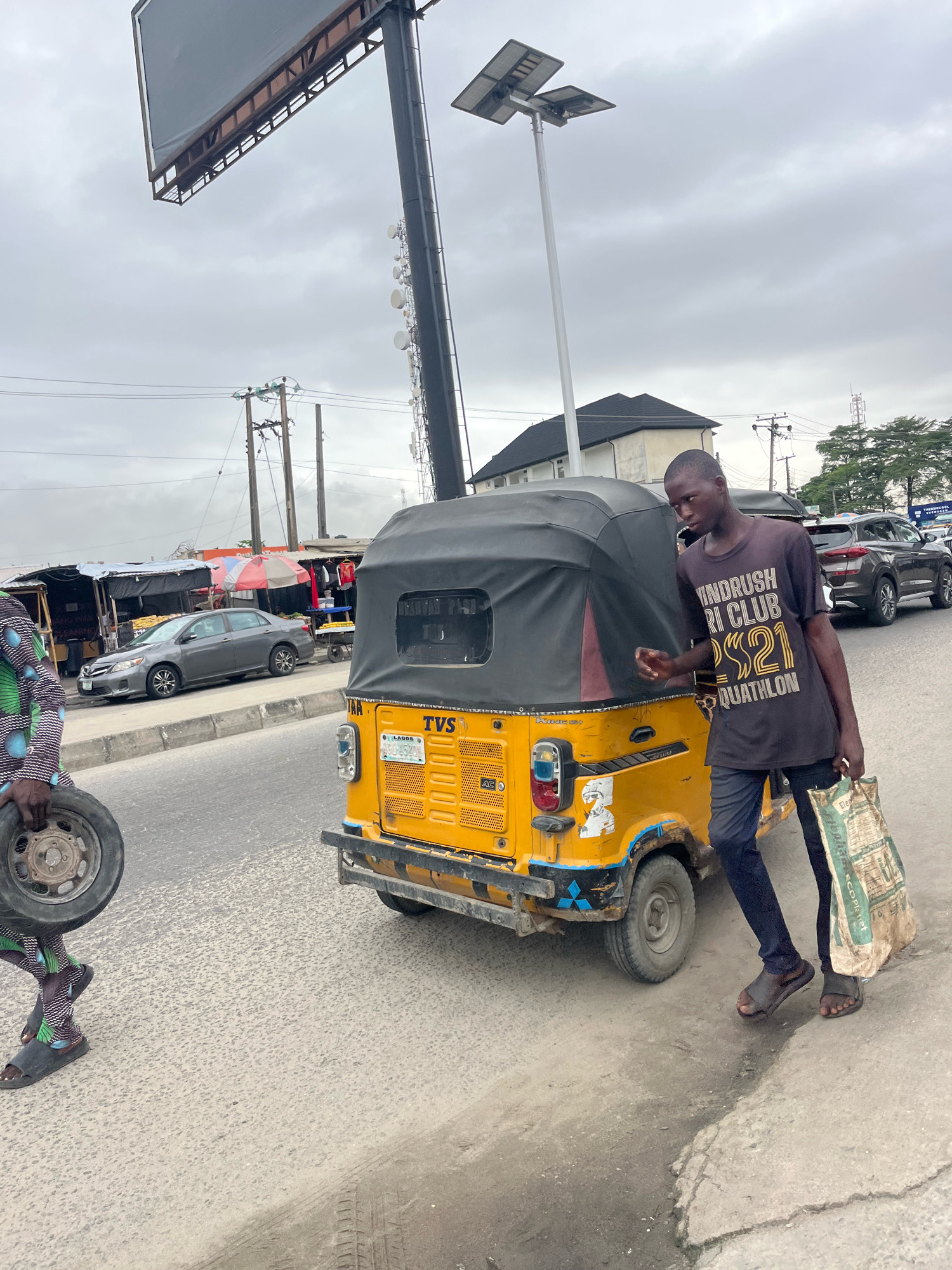 In Ado, Nigeria, on an overcast day, a young man stands beside a yellow auto-rickshaw, holding a large, woven bag in his right hand. He wears a dark t-shirt with white and gold lettering that reads "WINDRUSH TRI CLUB 2021 AQUATHLON," dark trousers, and dark sandals. His left hand rests on the side of the auto-rickshaw. The auto-rickshaw has a black canopy and a license plate from Lagos. To the left of the auto-rickshaw, another person, wearing a vibrantly patterned garment and dark sandals, carries a spare tire. In the background, several market stalls are visible, with awnings providing shade. A grey sedan is parked along the roadside, and further in the distance, other vehicles and trees can be seen. A tall pole with a large, blank billboard at the top and multiple antennas rises into the sky, accompanied by utility poles and wires, suggesting an urban or semi-urban setting.