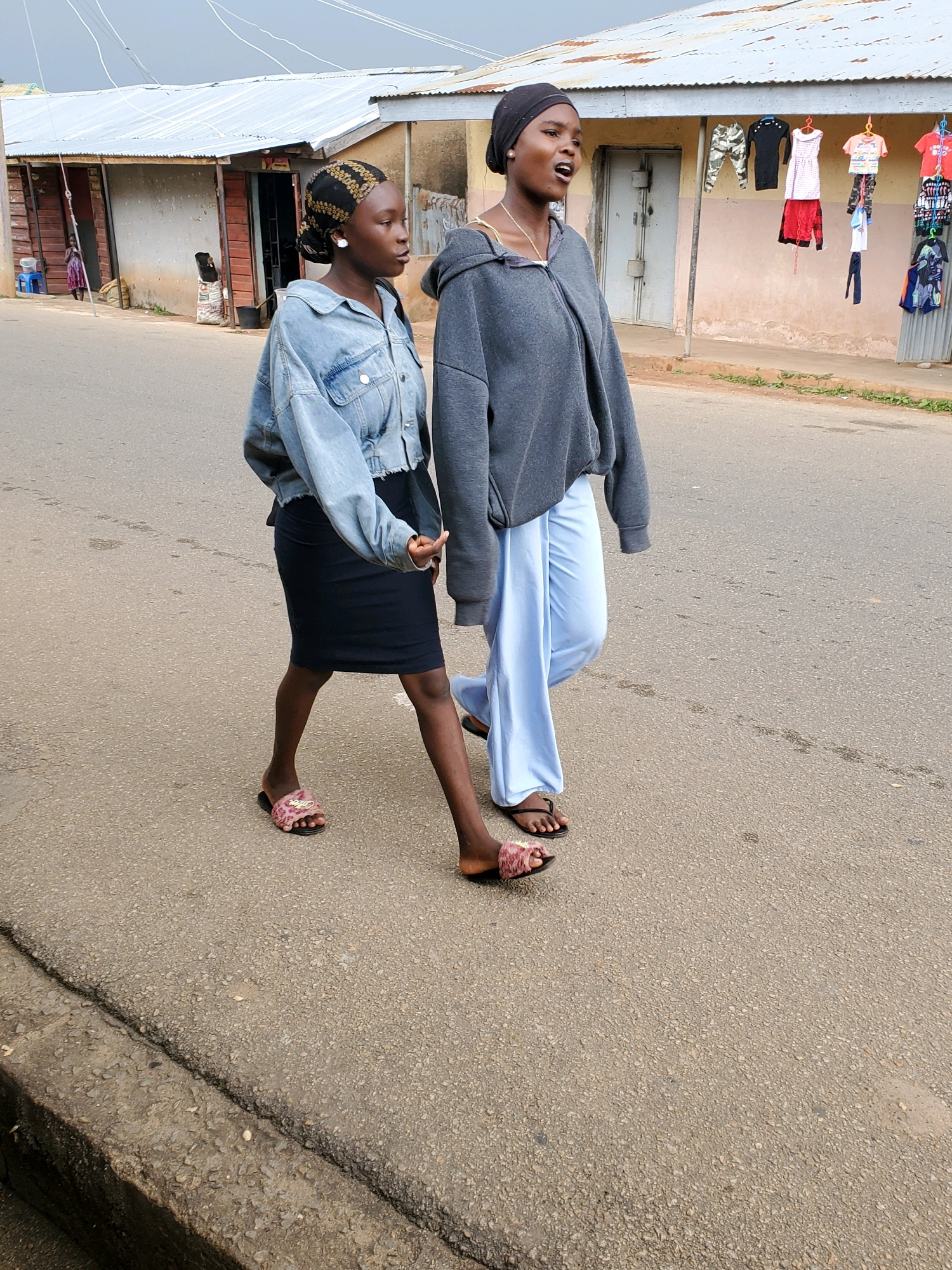 Two young women are walking on a paved street in front of a row of shops. The woman on the left is wearing a denim jacket, a black skirt, and patterned slides. The woman on the right is wearing a grey hooded sweatshirt and light blue wide-leg pants, with black sandals. The shops have corrugated metal roofs and wooden or metal doors and windows. Clothing items are displayed for sale on hangers in front of one of the shops. The sky is overcast. The location is identified as Jos, Nigeria.