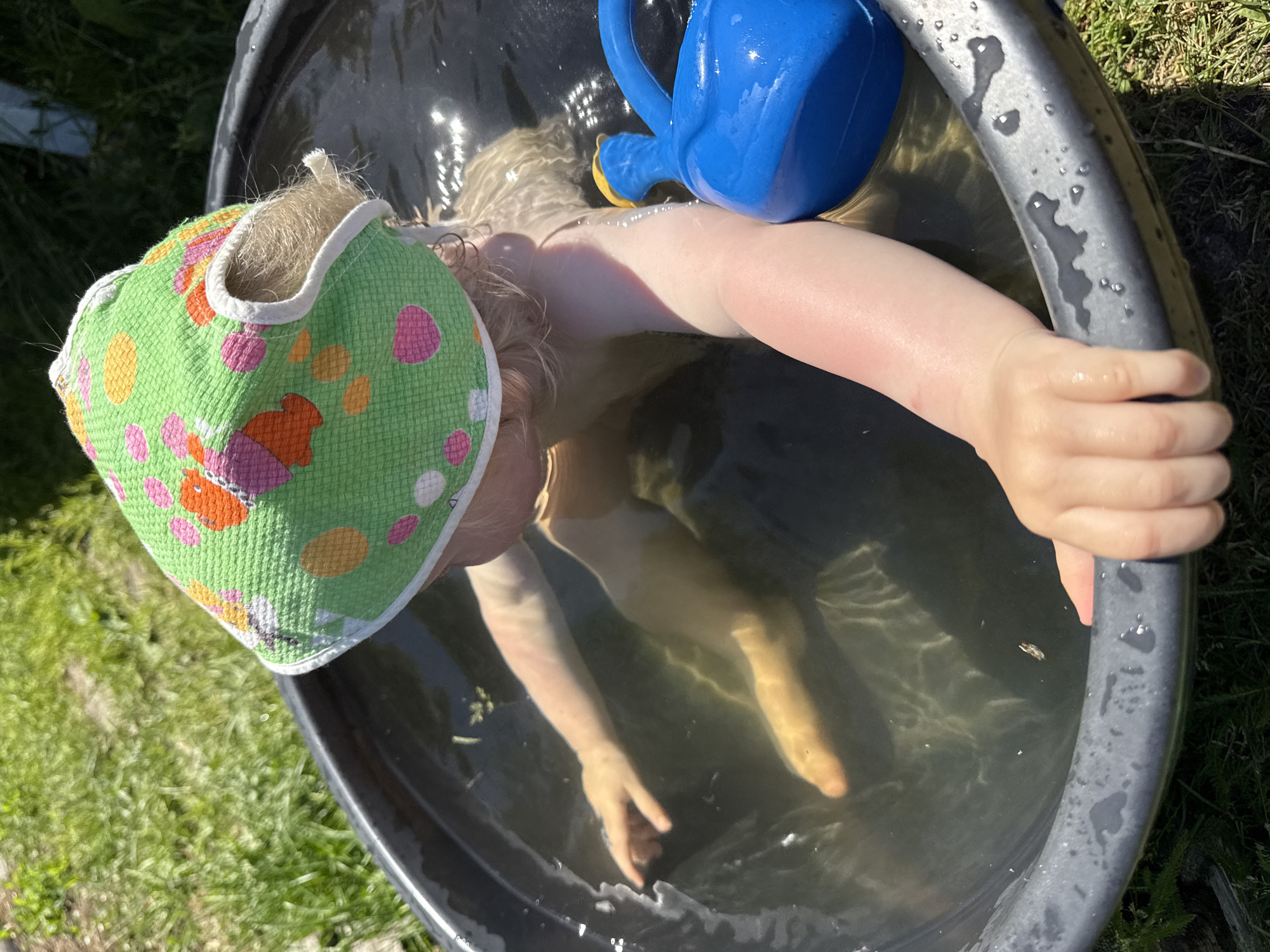 An overhead, slightly angled photograph captures a young child immersed in a round, dark-colored tub filled with water, positioned outdoors on a grassy surface. The child, observed from the torso up, wears a green sun hat adorned with multicolor polka dots and abstract patterns in pink, orange, and yellow, with a white brim. Blonde hair is visible beneath the hat and flows in the water around the child's head and neck. The child's arms and legs are submerged, with one arm resting on the tub's rim, fingers gripping the edge, and the other arm extending into the water. The visible skin on the child's upper right arm exhibits a distinct reddening, consistent with a mild sunburn. A bright blue plastic watering can with a yellow spout cap floats partially submerged near the child's right arm. The water in the tub is clear, reflecting bright sunlight that creates shimmering patterns and highlights small pieces of floating organic debris. The tub is surrounded by vibrant green grass, indicative of an outdoor, sunny environment.