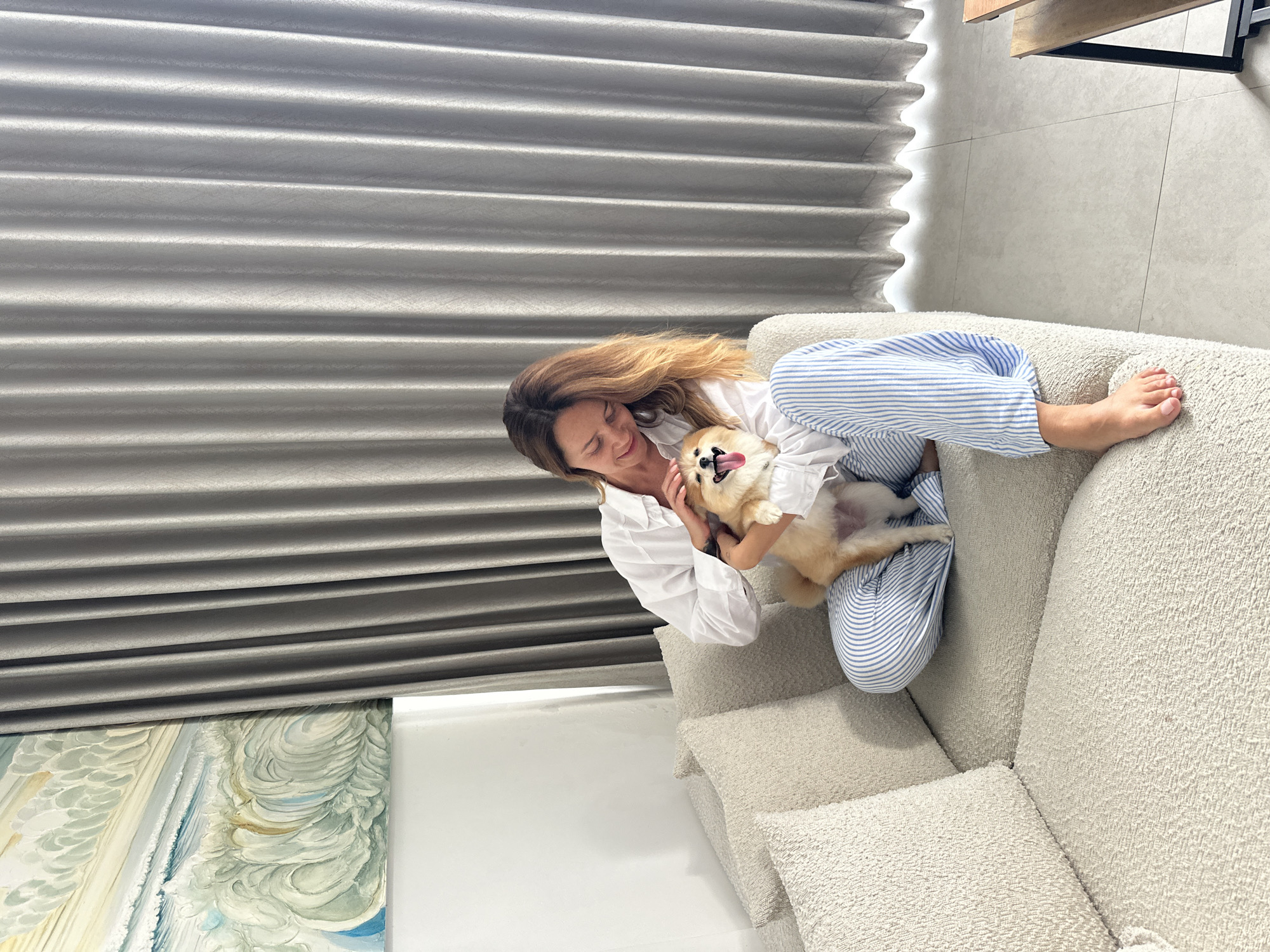 A woman with shoulder-length brown hair is sitting on a beige couch, holding a small, tan dog. The woman is wearing a white button-down shirt and blue and white striped pants. She is looking down at the dog and petting its head. The dog has its tongue out and appears to be happy. The couch has a textured fabric and is adorned with several throw pillows. Behind the couch, there are floor-to-ceiling gray curtains. To the left of the couch, a painting with blue and white abstract waves is visible on the wall. The floor is tiled in a light gray color. The overall scene suggests a comfortable and relaxed indoor setting.
