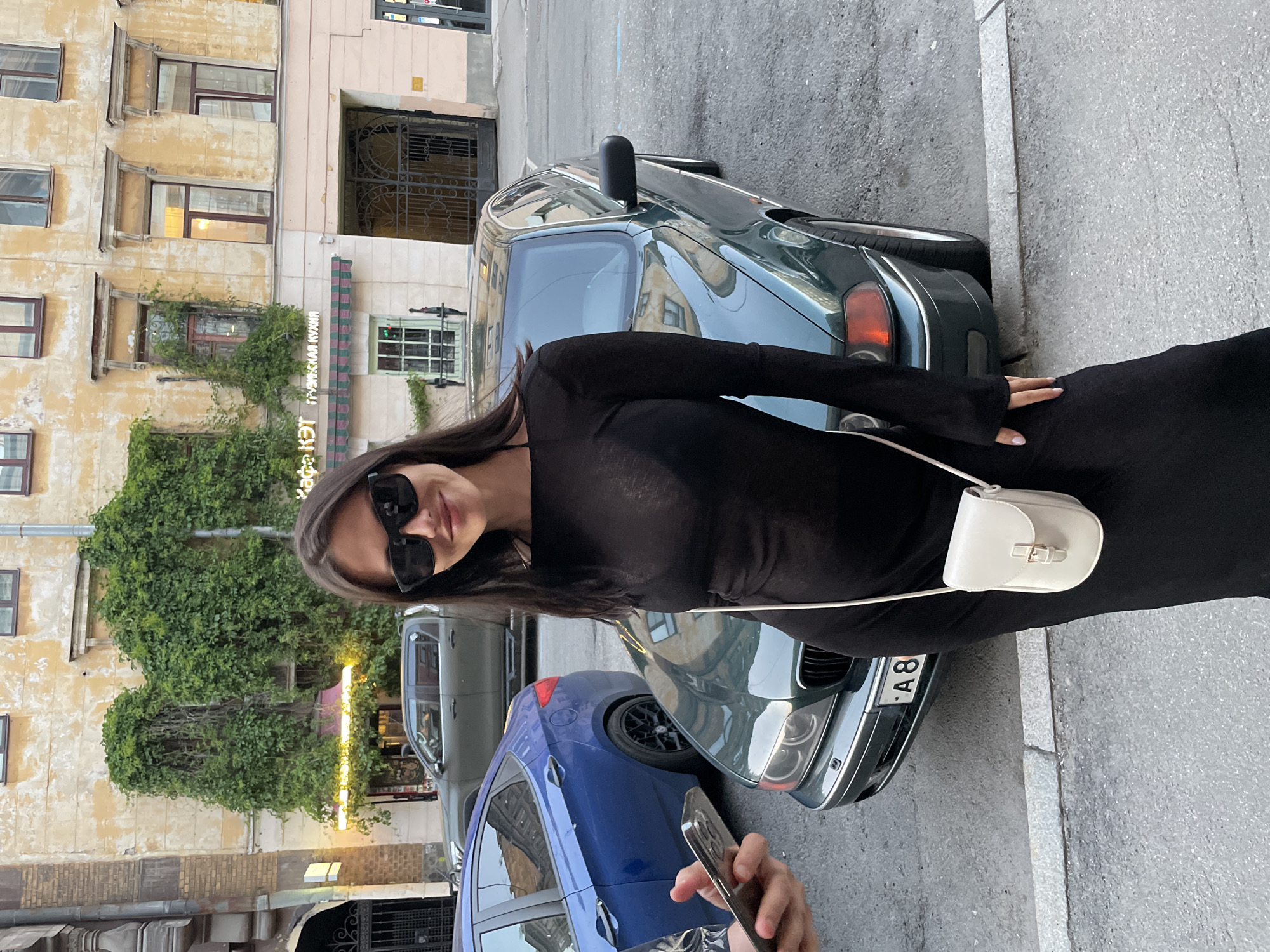 An outdoor, eye-level shot captures a young adult female posing on a sidewalk in Saint Petersburg, Russia, specifically on Sadovaya Ulitsa, in front of the "КАФЕ КЭТ ГРУЗИНСКАЯ КУХНЯ" (CAFE KET GEORGIAN CUISINE). The woman, with long dark hair, wears black sunglasses, a sheer long-sleeved black dress over a dark bra, and carries a small, off-white cross-body bag. Her left hand is visible, holding the bag strap; her nails have light polish. To her immediate left, another person's hand holds a silver smartphone, pointed towards her, indicating she is being photographed.
Behind the woman, a dark green BMW E39 sedan is parked on the street, displaying a partial Russian license plate with "А8" visible on the front. Further back and to the left, a blue sedan is partially visible, followed by a dark-colored pickup truck.
The background features a multi-story building with aged, yellow-ochre peeling paint, partially covered in dense green ivy, particularly around two upper-story windows. The ground floor of this building houses the "КАФЕ КЭТ ГРУЗИНСКАЯ КУХНЯ," identified by a sign above a striped green and red awning. The cafe's entrance area is illuminated with yellow lights. To the right of the cafe, a large arched entryway with dark, ornate metal gates leads into a building section with a light pink facade. The street is asphalt, separated from the concrete sidewalk by a curb. The overall lighting suggests late afternoon or early evening.