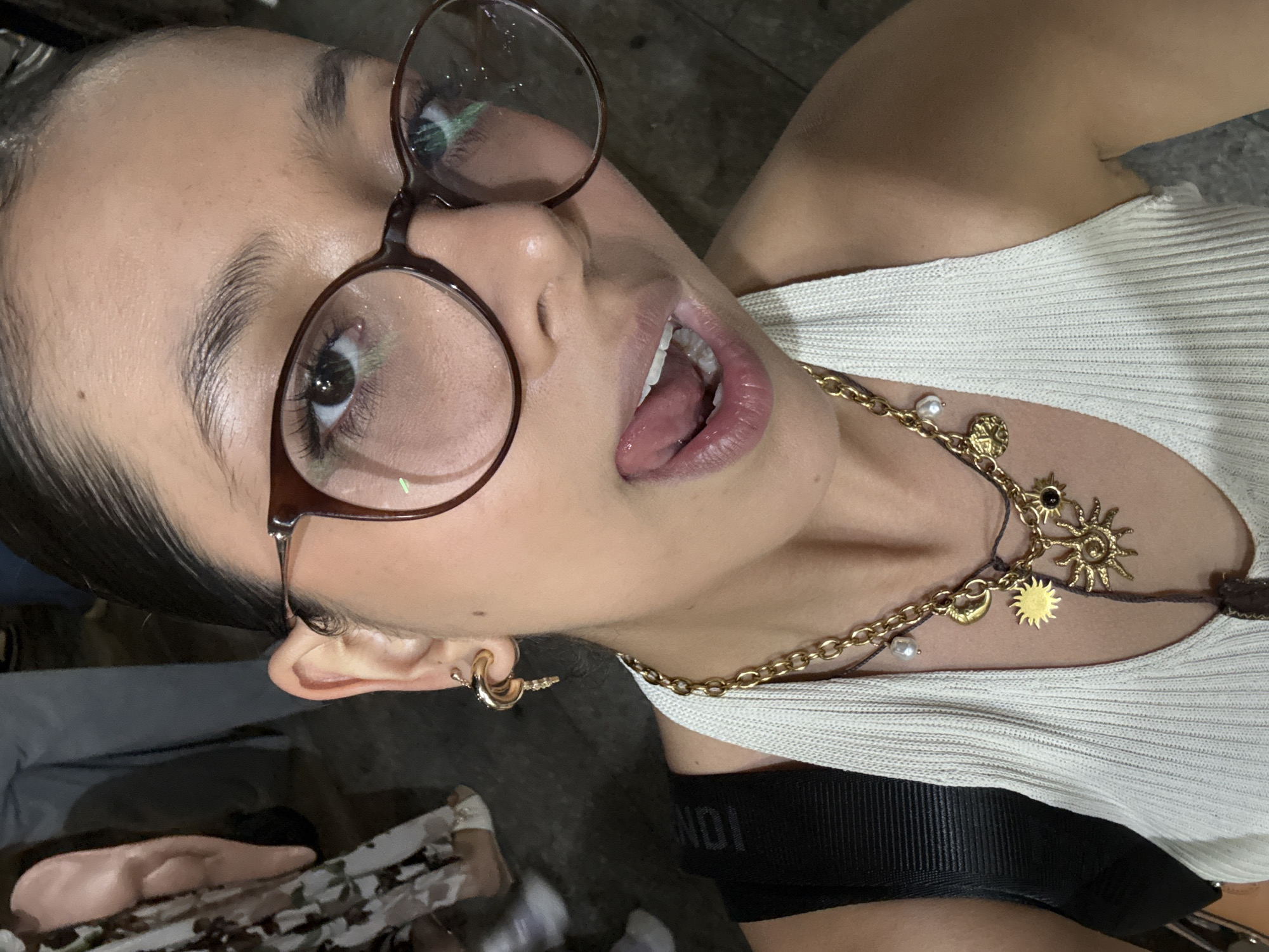 A close-up, tilted portrait shows a person with light skin, wearing glasses with a brown frame, and a gold chain necklace with charms. The person is wearing a white ribbed tank top over a black strap with the word "FENDI" visible. The person has gold hoop earrings and is sticking out their tongue. The background is blurry, suggesting an outdoor setting, potentially in the city of Veracruz City, Mexico.