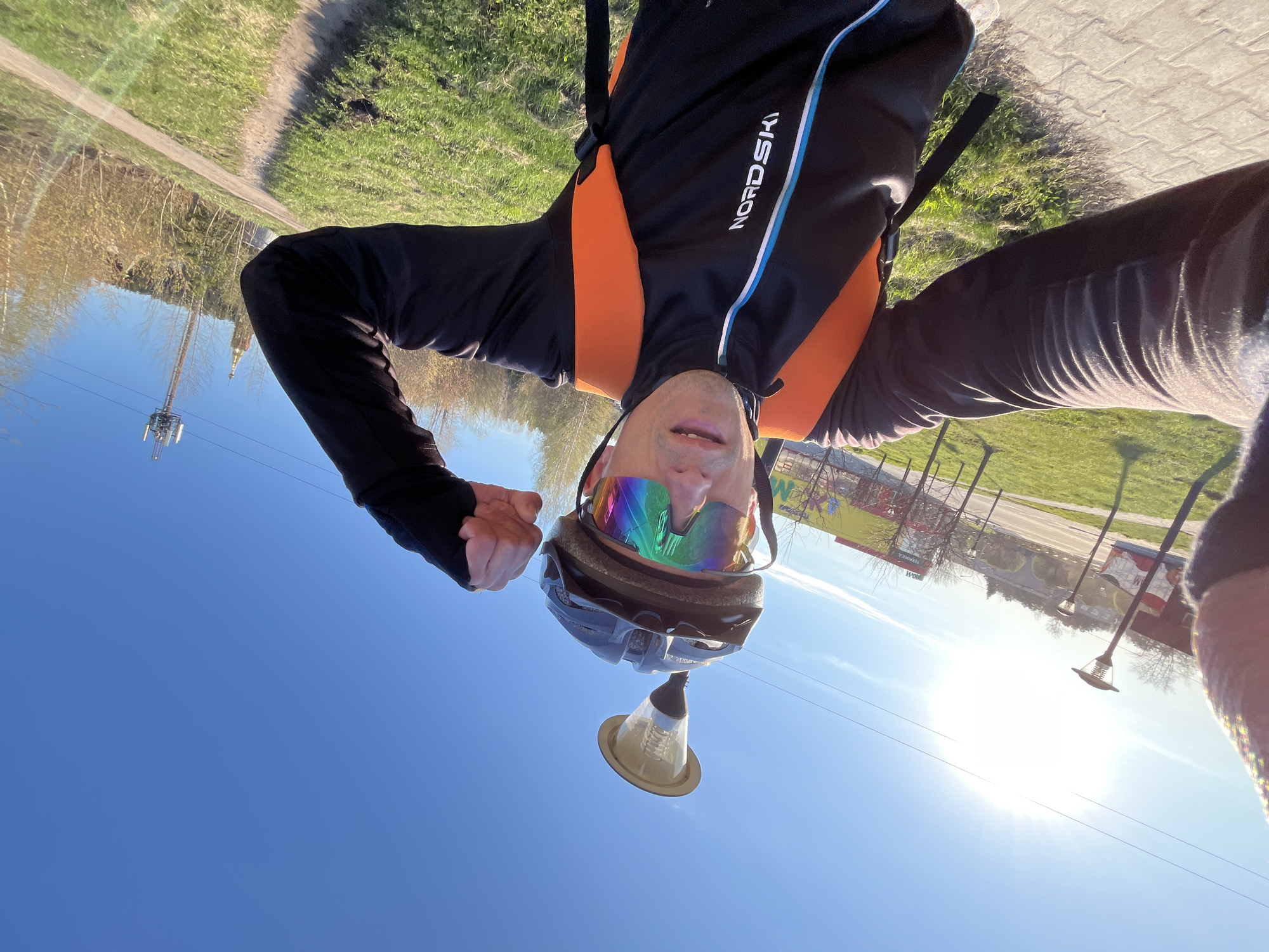 The image is a selfie taken by a person wearing a black jacket with "NORDSKI" printed on the sleeve, orange straps, colorful sunglasses, and a helmet with an attached light fixture. The person is standing outside on a sunny day with a clear blue sky. In the background, there is a grassy area, a paved path, trees, a cell tower, and buildings. Based on the information provided, the location is Chaikovsky, Russia. There are overhead power lines visible in the lower part of the frame. The image is rotated.