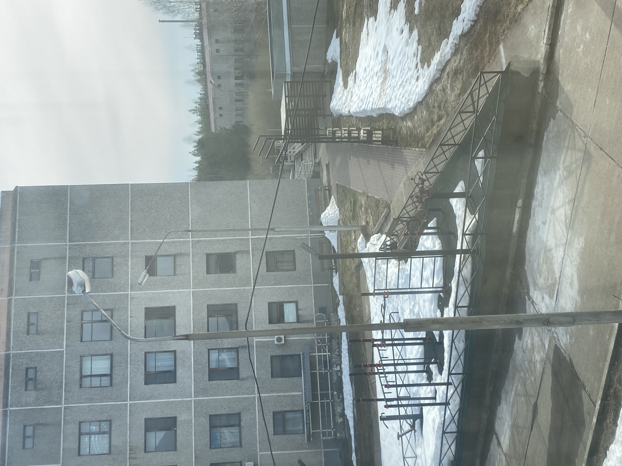 A drab, gray, post-Soviet apartment building dominates the scene, its facade punctuated by rows of windows. A bare street lamp extends from the building. In front of the building, a series of metal structures are visible, possibly scaffolding or a framework for construction. Patches of melting snow are scattered on the ground, intermixed with areas of dry grass. In the background, other buildings and trees are visible, blurred by distance. The overall scene appears to be cold and overcast. It is located in the city of Марковский, Russia.