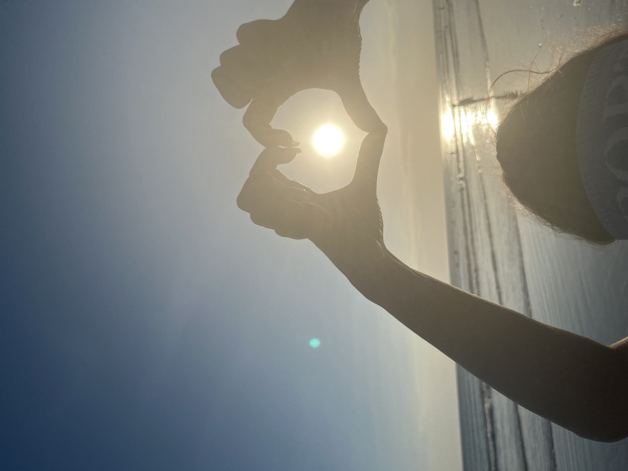 The image shows a person's hands forming a heart shape around the sun, silhouetted against a bright sky. The sun is the focal point, positioned in the center of the heart. The background is a blend of clear blue sky and a hazy, bright horizon. To the right, there is a suggestion of water, likely the ocean, with the sun's reflection shimmering on its surface. A person's head and shoulder, also in silhouette, are visible on the far right. The lighting indicates it is either sunrise or sunset. The overall mood is peaceful and serene.