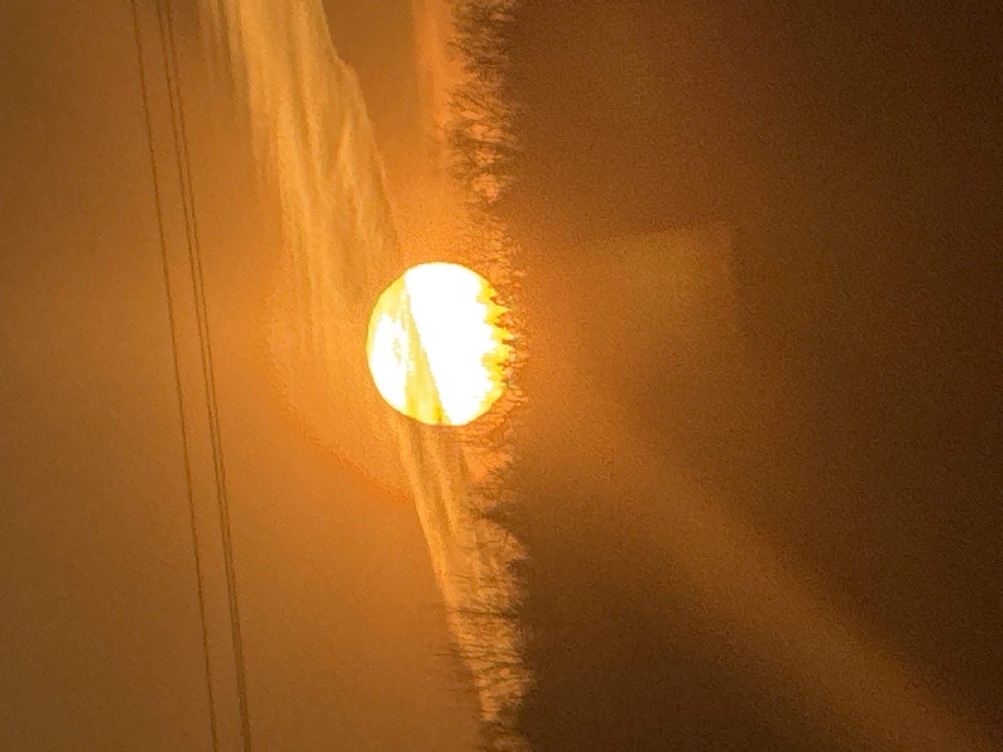 The image shows the sun partially obscured by haze or smoke, casting an orange glow across the sky. Power lines are visible in the left portion of the frame. What appears to be a treeline or dense vegetation is silhouetted against the sun's light. The location is unknown and is not possible to confirm that it is Chaikovsky, Russia based on the information within the image.