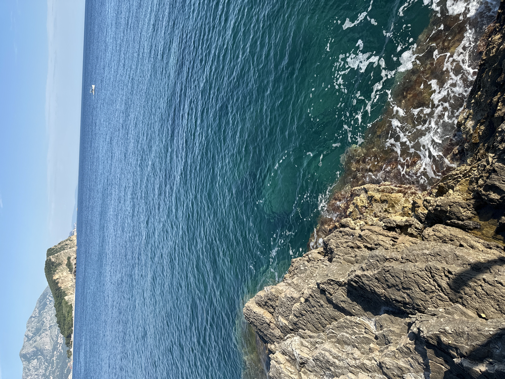The image shows a view of the Adriatic Sea from a rocky cliff near Budva, Montenegro. The water is a deep blue color, with white foam near the rocks. The cliff is made of layered, jagged rocks. In the distance, a cliff covered with greenery and a sliver of bright blue sky can be seen. There is a small white boat in the distance.