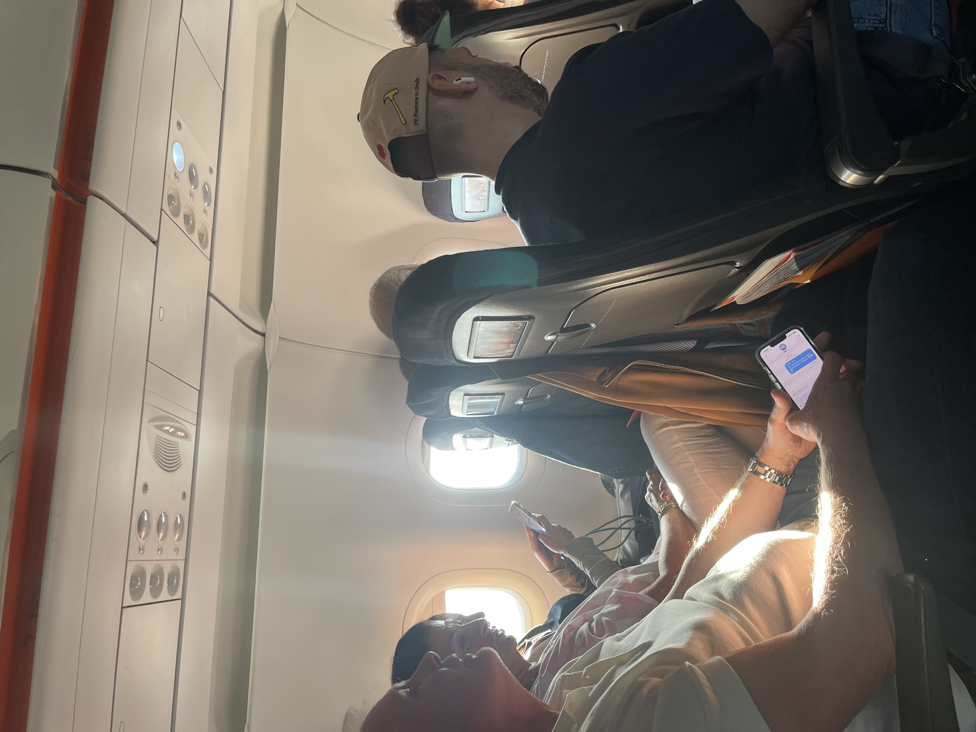 The image shows the interior of an airplane cabin during daytime. Several passengers are visible in their seats. One person is holding a smartphone, presumably interacting with it. Another passenger in the foreground is wearing a white t-shirt, while a man behind him is wearing a cap with a hammer on it. The scene is bright, illuminated by sunlight entering through the windows. The location is most likely the Lyon–Saint-Exupéry Airport in Colombier-Saugnieu, France.