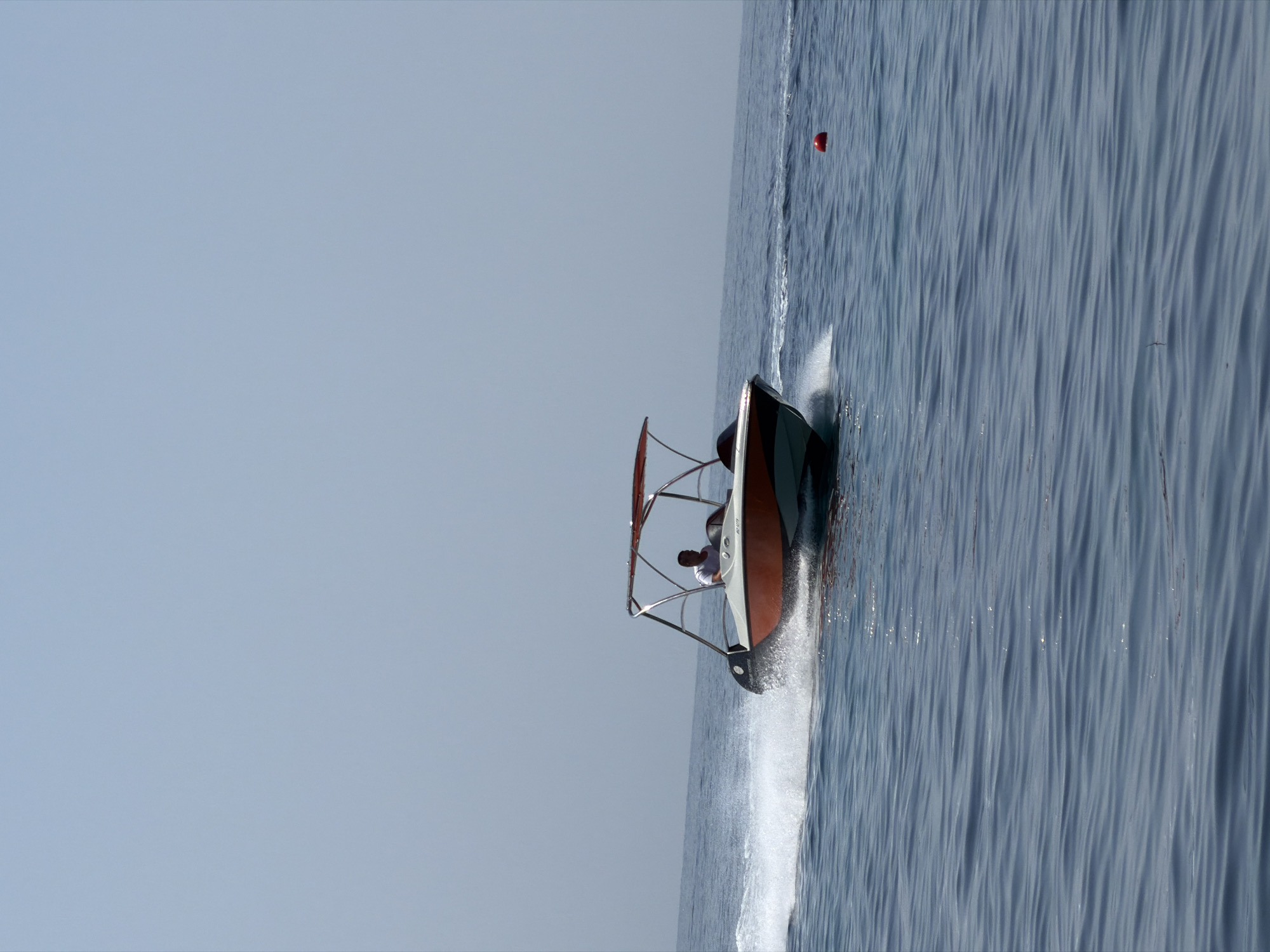 A man in a white t-shirt is piloting a small, orange and white motorboat across calm blue water. The boat is moving at a moderate speed, creating a white wake behind it. The boat has a canopy supported by metal poles. In the distance, the sky is a hazy, pale blue, and the horizon is a clear line between the water and the sky. A single red buoy is visible floating on the water to the right of the boat. The scene appears to be taking place in an open body of water, possibly the Adriatic Sea near Budva, Montenegro, given the characteristic blue water and clear skies often seen in the region.