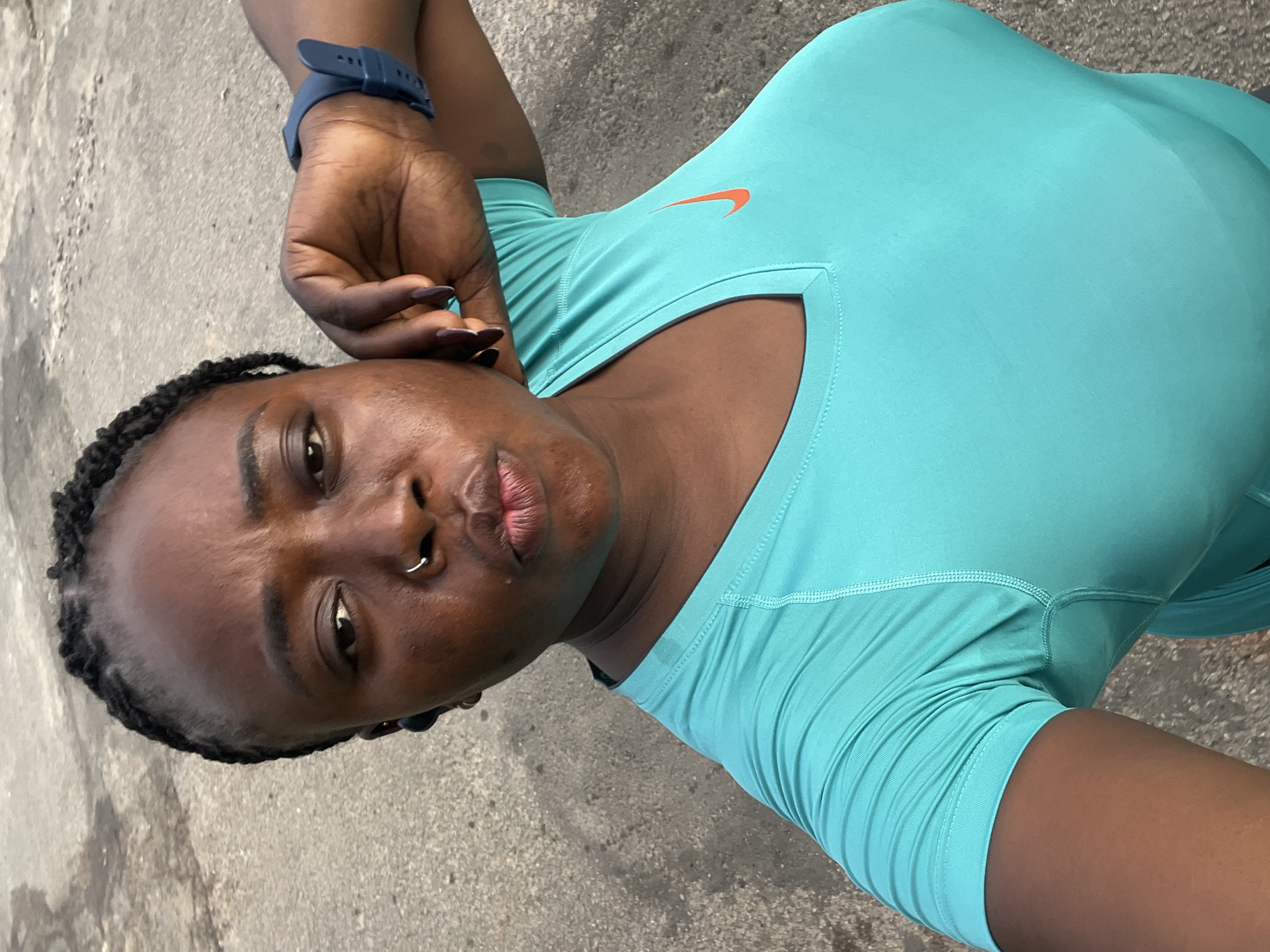 A medium shot shows a Black woman from the chest up, outdoors on a paved surface. She is wearing a turquoise v-neck t-shirt with an orange Nike logo on the left side of her chest. Her left arm is bent at the elbow, and her hand is resting on her head, with her fingers splayed. She has dark braided hair, and she is wearing a silver nose ring. She has dark nail polish on her fingernails. A dark blue watch is visible on her left wrist. The woman is looking down and slightly to her left. The background is a grayish, textured surface, possibly asphalt or concrete, indicative of a sidewalk or road in Abuja, Nigeria.