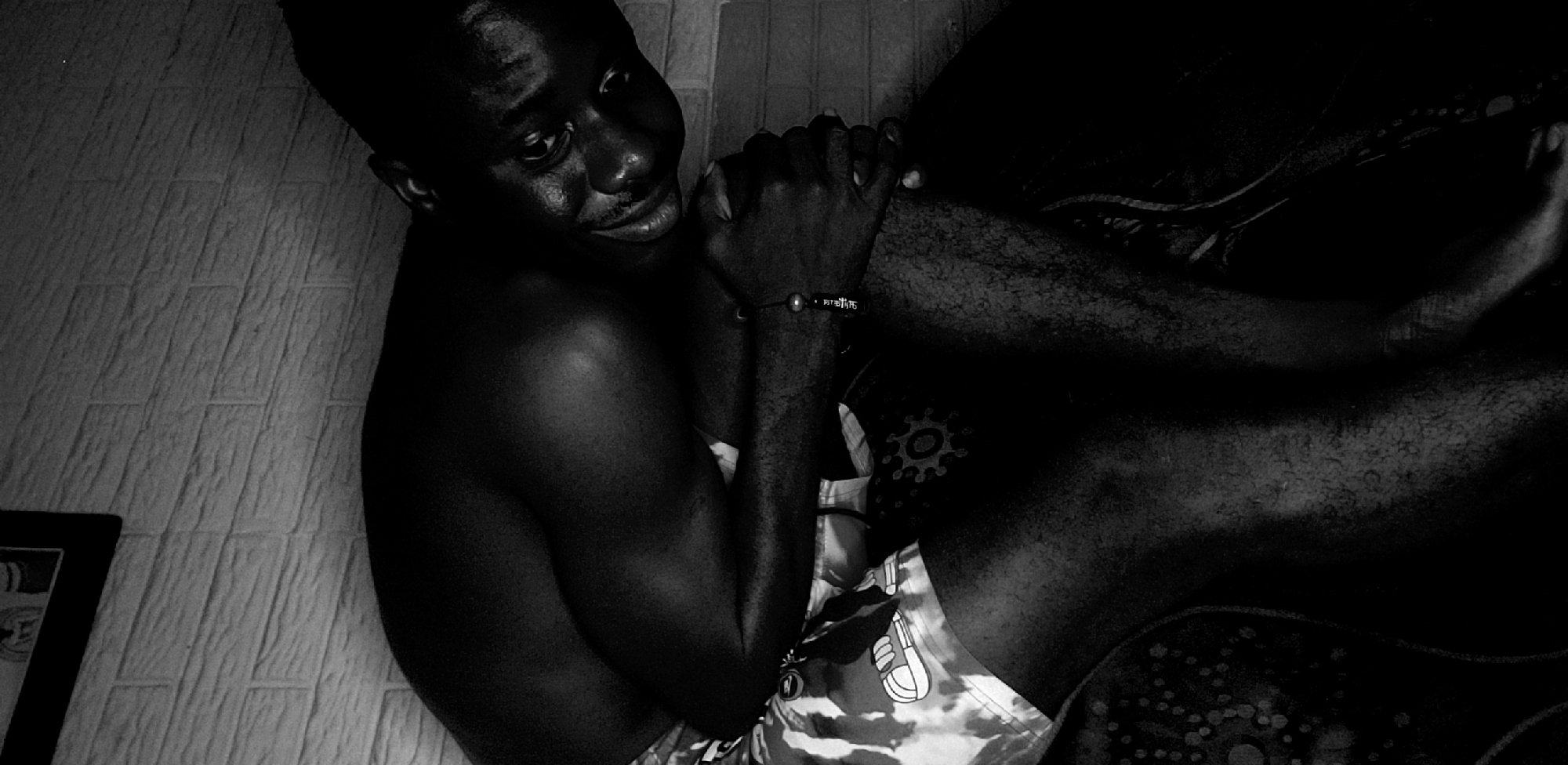 A Black male is sitting on a bed in a room with off-white brick-patterned walls. The male is wearing patterned boxer shorts and a bracelet. He has dark skin and short hair. His right arm is positioned over his left arm, his hands clasped together. He is looking to the right of the frame. The room is dimly lit, with a dark bed and patterned sheets. A picture frame is in the lower left corner of the image. The location is Narayi, Nigeria.