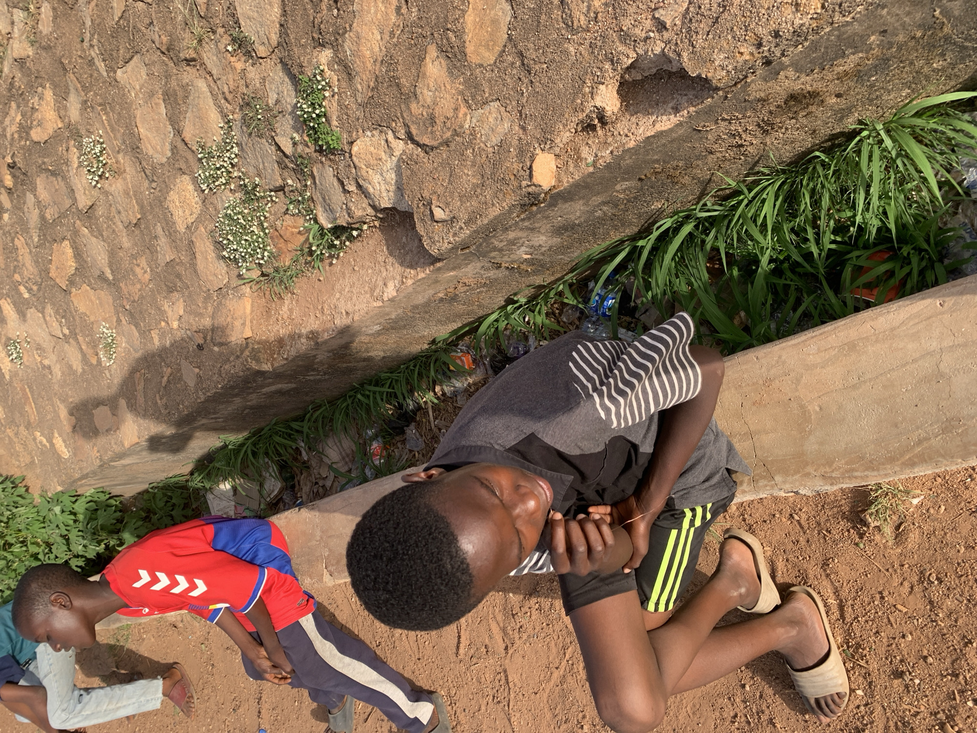 In Gura Top, Nigeria, two young people are near a stone wall. One youth wears a red shirt with white chevrons and blue shoulder and back panels paired with navy trousers that have white stripes. The other youth wears a gray and black striped shirt and black shorts with yellow stripes and neutral colored sandals. The stone wall has green vegetation, and some trash is caught in it.