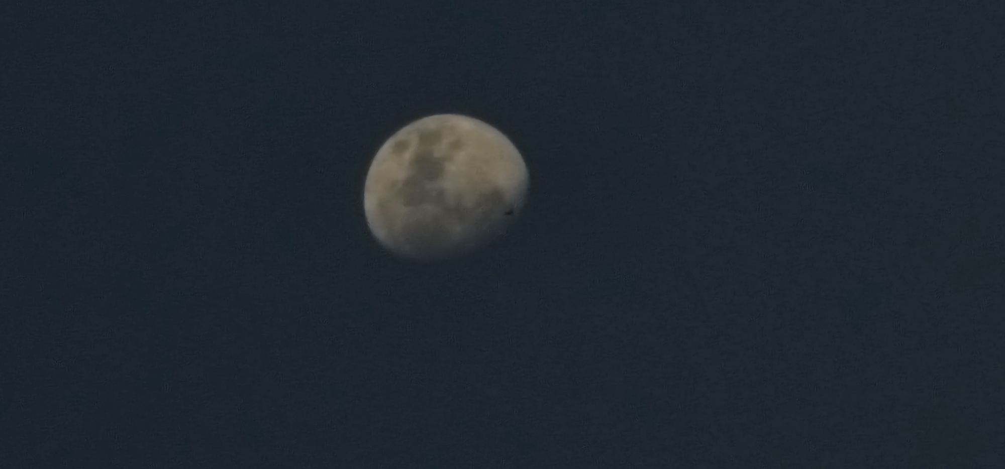 A crescent moon is visible in a dark sky. The moon is illuminated from the right, and its surface shows visible craters and maria. A dark, silhouetted object, possibly an airplane or bird, is in transit in front of the moon. The background is a deep blue, suggesting nighttime or twilight. The image is taken from an unknown location.