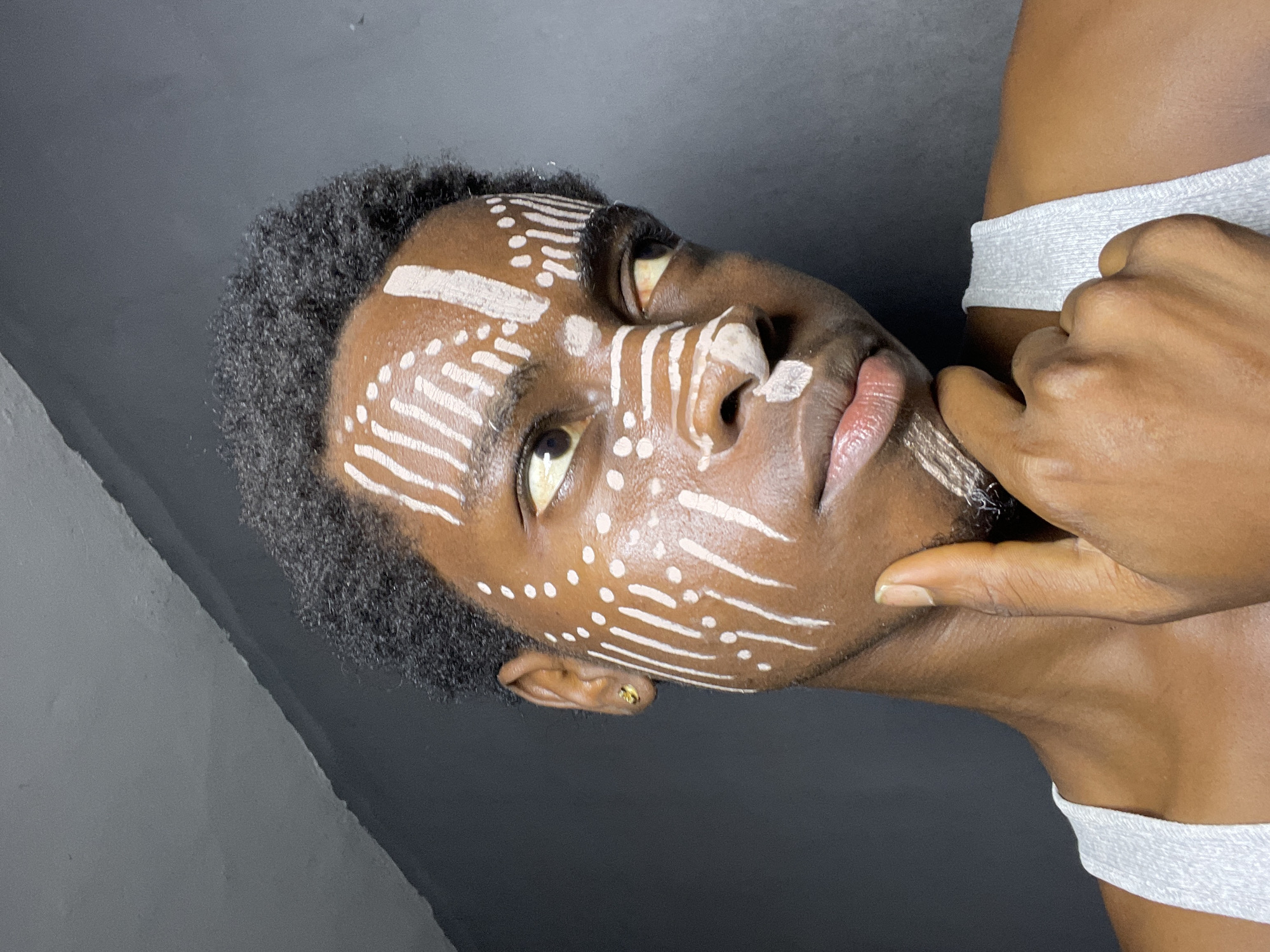An African man with dark brown skin is shown in a close-up portrait. He has short, dark, kinky hair and is wearing white face paint in lines and dots. He is touching his chin with his right hand and is wearing a white top. The background is a dark grey. It is not possible to identify if this image was taken in Nkpor, Nigeria.