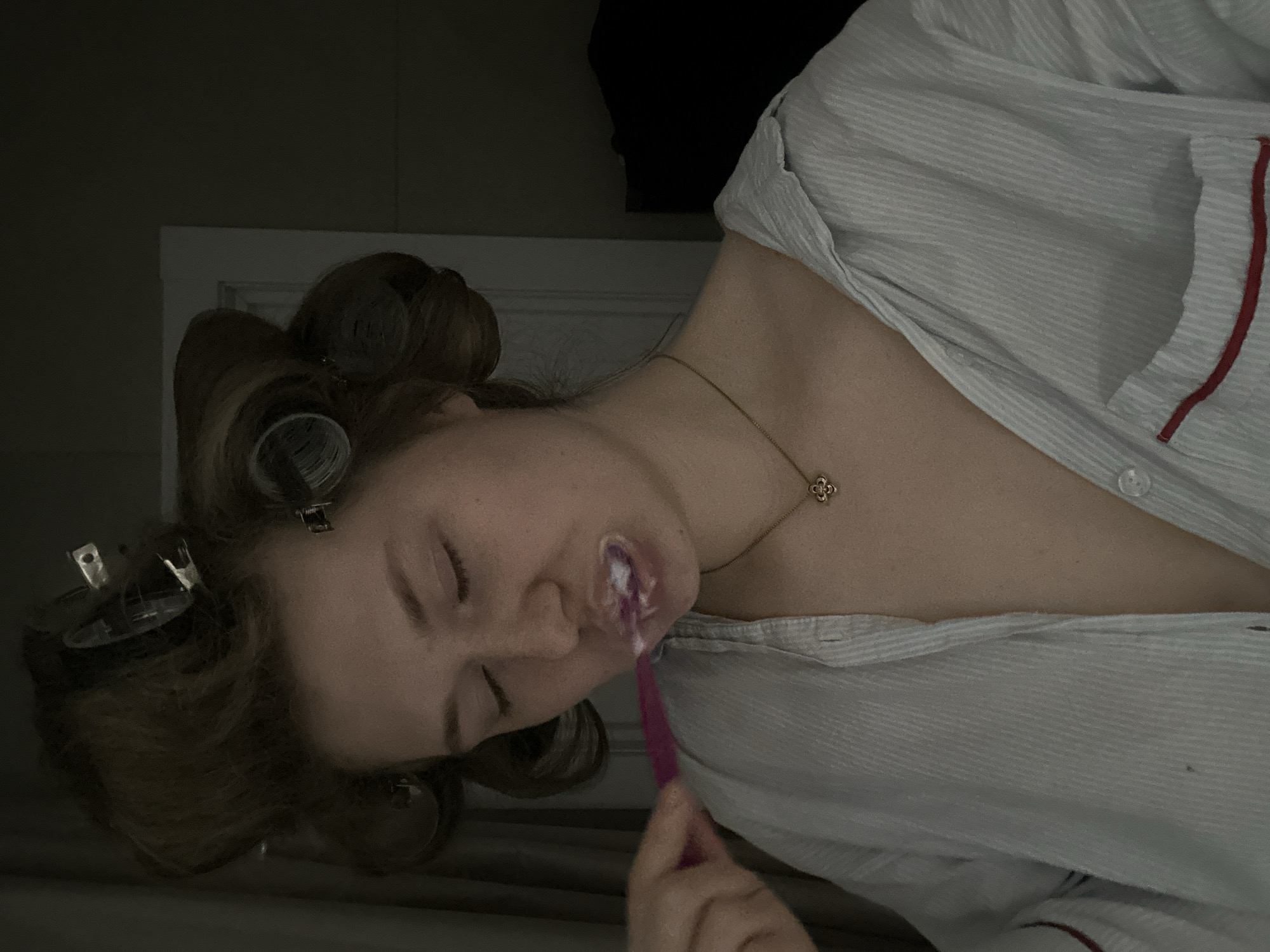 A woman is brushing her teeth while lying down. She has curlers in her hair. She is wearing a light blue striped pajama top with red trim. She is wearing a gold necklace. The background is dark and includes a white cabinet. The location is indeterminable based on the image alone. The city of Kazan, Russia is not evident in this image.