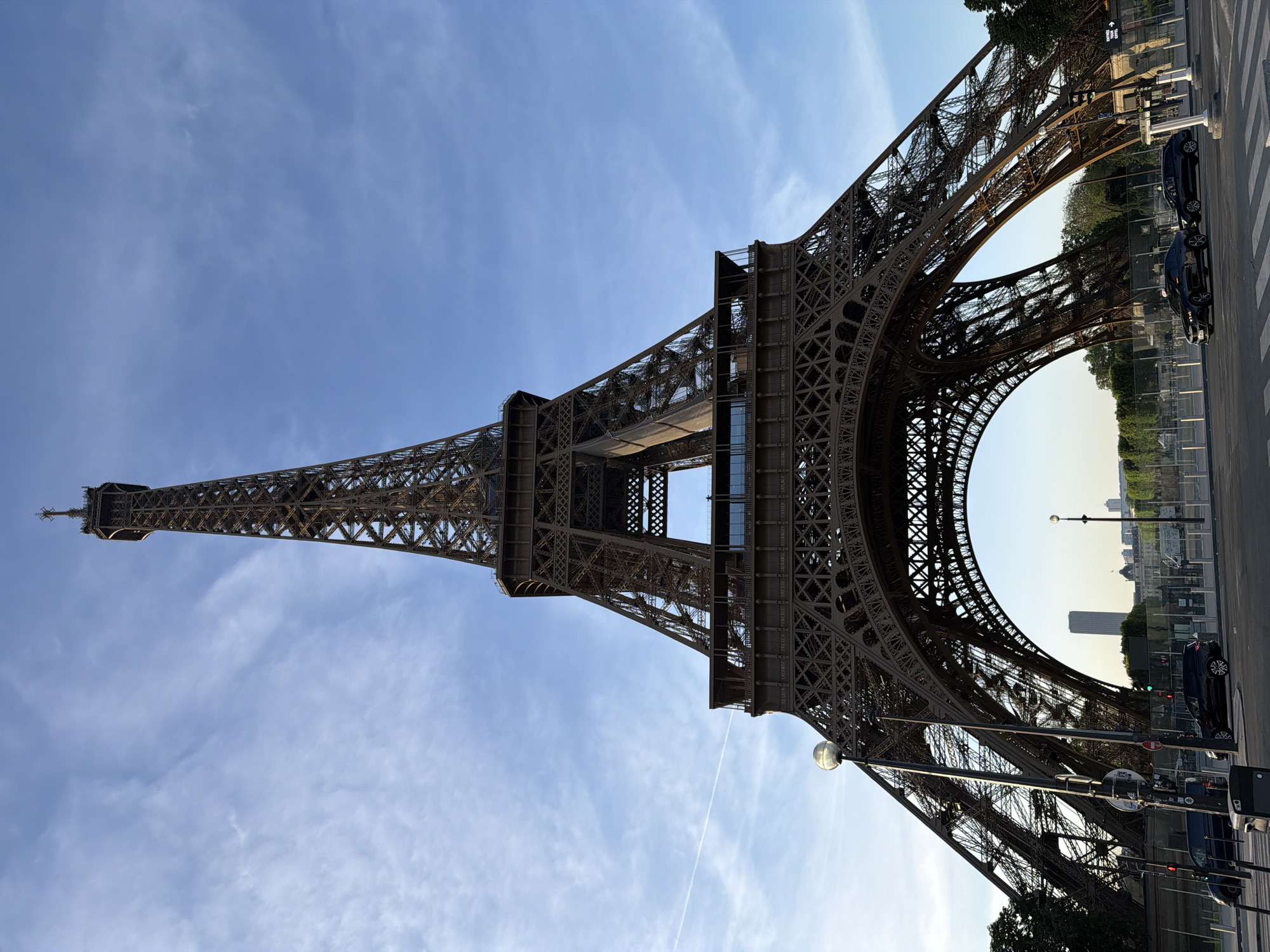 The image shows the Eiffel Tower in Paris, France, oriented vertically. The sky is visible behind the tower, with light blue hues and wisps of clouds. The tower's intricate metal latticework is prominent. On the right side of the image, at the base of the tower, are trees, streetlights, buildings in the distance, and two parked cars. The scene appears to be captured during the daytime.