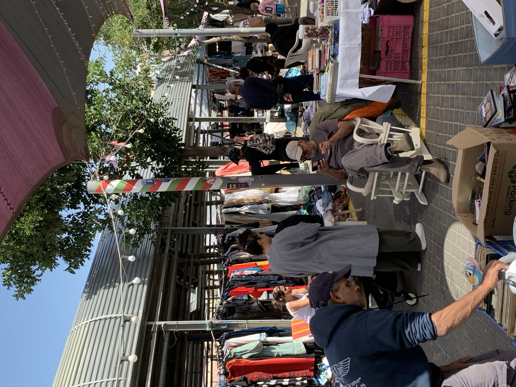The image shows a bustling scene at a flea market in Tel Aviv, Israel. People browse through stalls laden with second-hand clothing and various goods.
An older man in a blue hoodie and a baseball cap is sitting and looking at items in a cardboard box. Another man is sitting on a white plastic chair, looking at his cell phone. Other shoppers are standing behind the stalls. The market is partially shaded by a corrugated metal roof and canvas awnings. Decorations, like strings of lights and colorful pennants, hang overhead. The ground is paved with grey brick.