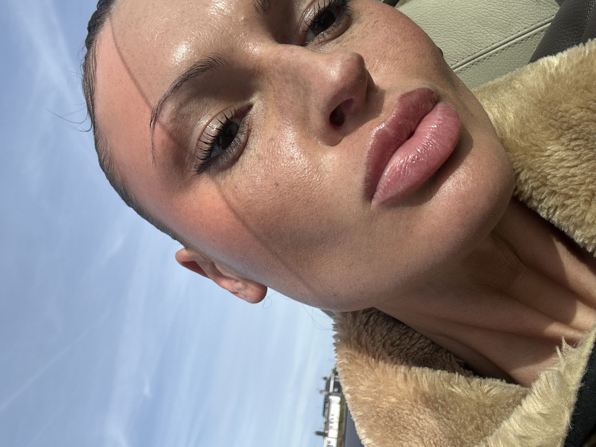 A close-up, low-angle selfie photograph of a young woman's face. The woman has fair skin with visible pores, dark eyes, and full, glossy lips. She is wearing minimal makeup. Her dark hair is pulled back, and there are stray hairs visible on her forehead. A portion of a tan-colored furry jacket or collar is visible on the right side of the frame. The background shows a pale blue sky with faint cloud patterns. In the distance, near the bottom of the frame, are blurred rooftops of buildings. Based on the style of the buildings and the sky conditions, and combined with the fur-lined coat, it is likely that the photo was taken outdoors in the Folkestone and Hythe District, United Kingdom.