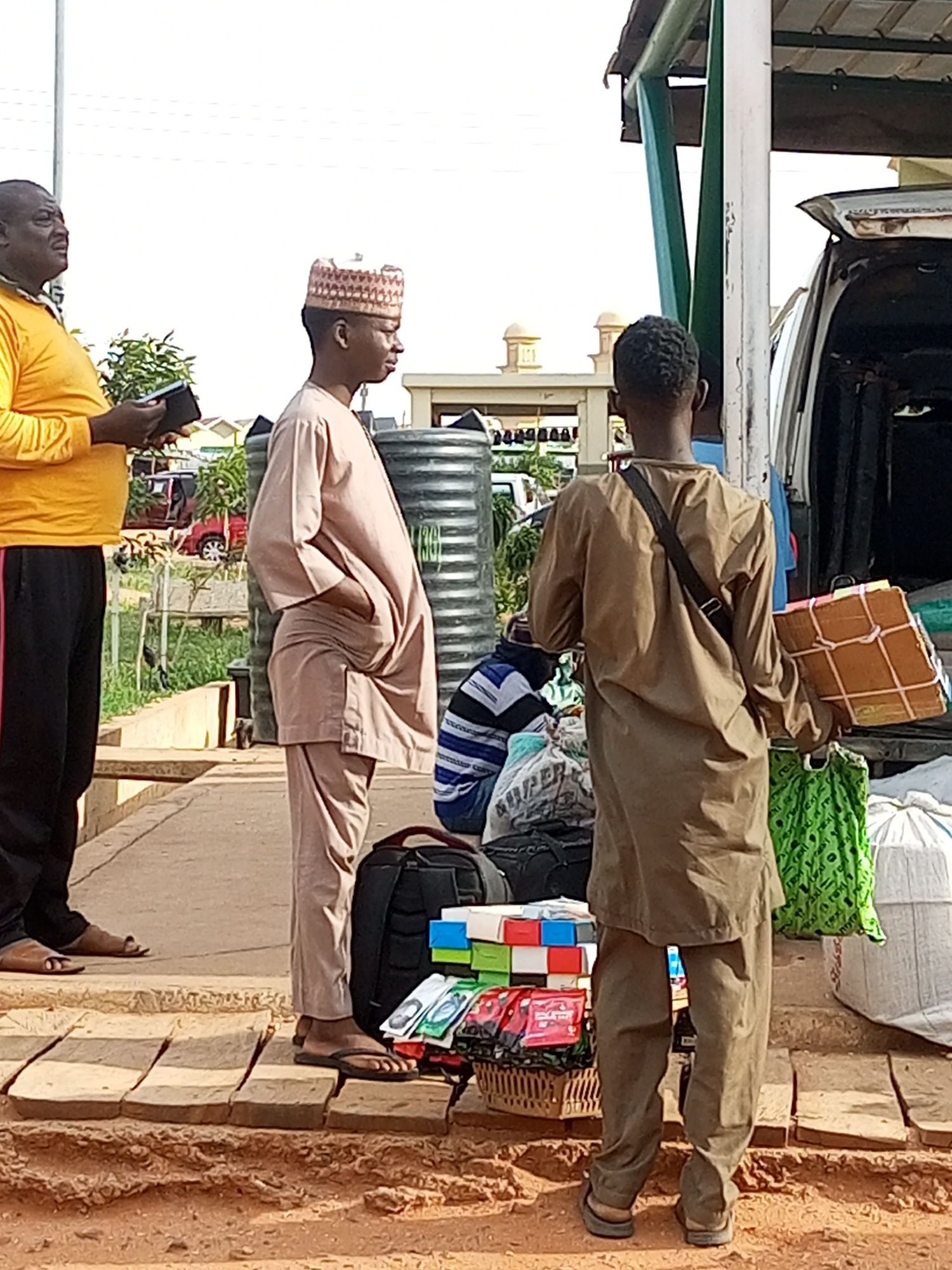 In the city of Gombe, Nigeria, three men are engaged in activity near a vehicle. The man on the left, wearing a yellow shirt and black pants, holds a black object, possibly a phone or wallet, and looks to the right. In the center, a man in a light brown, traditional outfit and a patterned cap stands with his hands in his pockets, facing away from the camera. To the right, a third man in a brown, traditional outfit stands with a brown box wrapped in string in his right hand and a green bag over his left shoulder. He is facing away from the camera, towards the open trunk of a white vehicle. In front of him, a collection of items are displayed, including a basket of packaged goods and several white and green bags of varying sizes. The scene appears to be outdoors, with a dusty ground and what looks like a raised concrete walkway. In the background, there are buildings with domed roofs and some greenery.