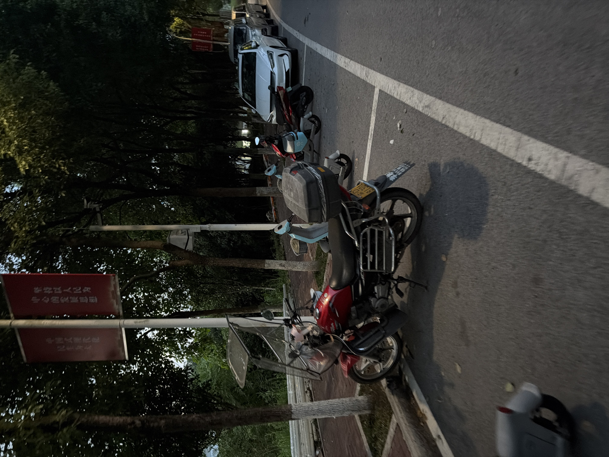 The image depicts a street scene in Jiangjin, China, with a focus on parked vehicles and surrounding environment. In the foreground, a red motorcycle with a clear windshield and a black storage box on the rear is parked along the curb. To its left, a light gray electric scooter is partially visible. Further down the street, several other vehicles are parked, including a white sedan, a silver SUV, and other motorcycles. The street is lined with trees, and banners are visible hanging from poles. The lighting suggests it is either early morning or late evening, with shadows cast by the trees. The ground is paved, and white lines delineate parking spaces. The overall scene is quiet and still, with no people visible.