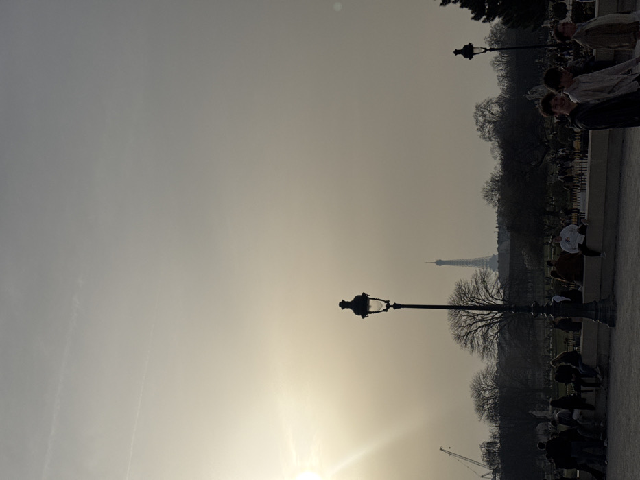 A photograph captures a hazy, sun-drenched outdoor scene in a park in Paris, France, likely during late afternoon or early evening. The sun, positioned in the upper-left quadrant, emits a strong flare, backlighting much of the foreground and midground. The sky above is a pale, hazy grey with faint contrails.
In the center-left foreground, a tall, ornate black street lamp stands vertically, its details visible despite the backlighting. Below this lamp, a low, light-colored stone wall or ledge extends horizontally across the frame. Approximately ten individuals are seated or standing casually along this ledge on the left side, mostly silhouetted. To the right of the central lamp, another similar street lamp is partially visible. Three young adult males walk towards the right side of the frame in the immediate foreground; one looks towards the camera with a faint smile, while the others face forward. Their attire is casual.
The midground features a public park setting with numerous bare deciduous trees, indicating late winter or early spring. Various individuals are scattered throughout the park's pathways and green spaces. In the background, the distinctive silhouette of the Eiffel Tower rises above the distant tree line, slightly obscured by atmospheric haze. A large construction crane is visible on the far left horizon, partially obscured by the sun's glare. The overall environment appears to be a populated urban park.