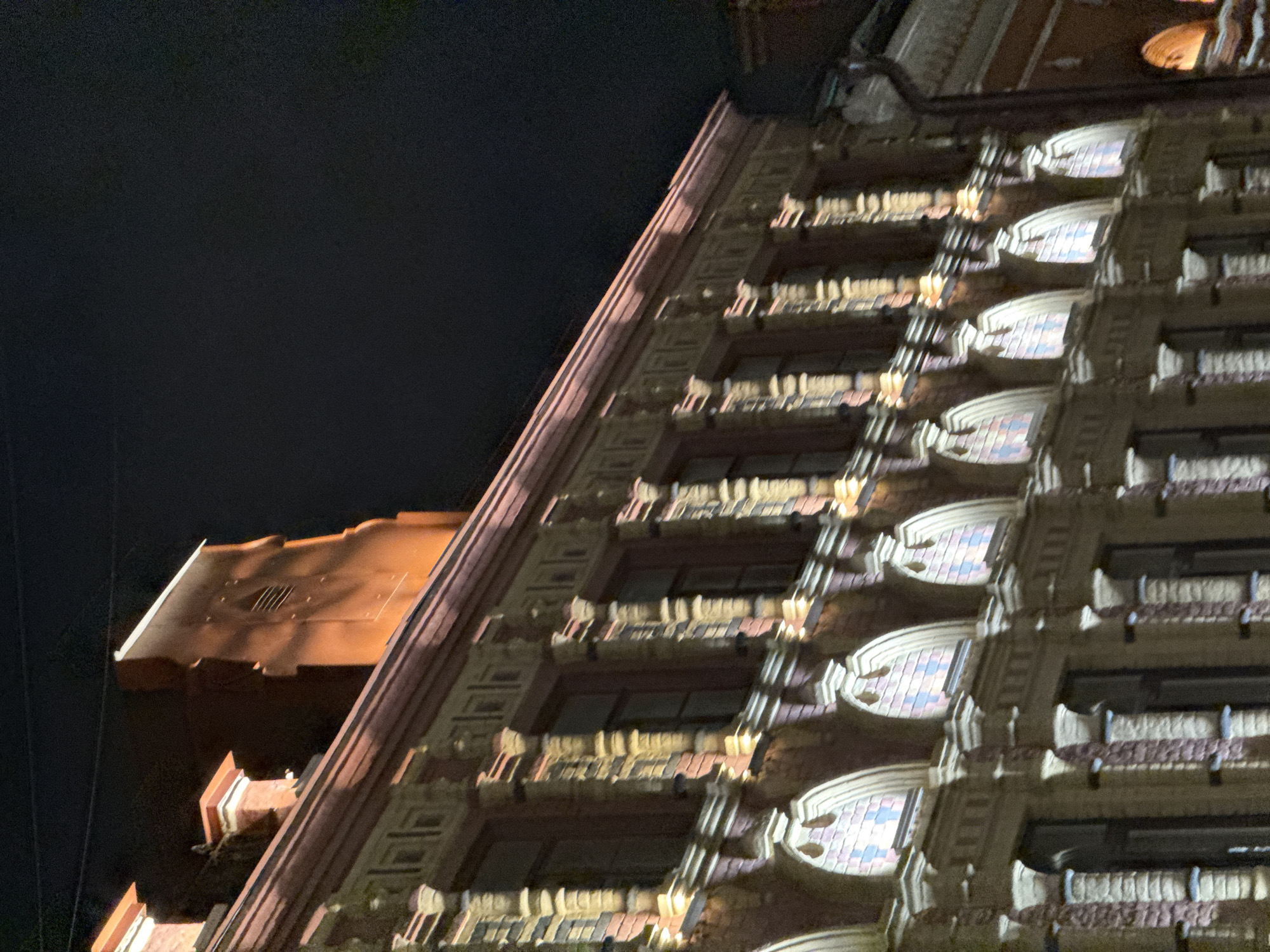 The image shows the facade of an ornate building at night, illuminated by artificial lights. The building features intricate architectural details, including decorative cornices, columns, and arched windows with stained glass. The overall style appears to be historical, possibly from the late 19th or early 20th century. The building is situated in a city environment, as suggested by the presence of what looks like a street or alleyway to the right. The sky is dark, indicating it is nighttime. The lighting casts warm, yellow and orange tones on the building's facade, highlighting its textures and decorative elements. No people are visible in the image. The building's architecture, particularly the style of the windows and decorative elements, is reminiscent of buildings found in historical city centers. Considering the context of the provided crops, which offer closer views of the building's details and a glimpse of neighboring structures, it is highly probable that this scene is located in the historical center of Moscow, Russia.