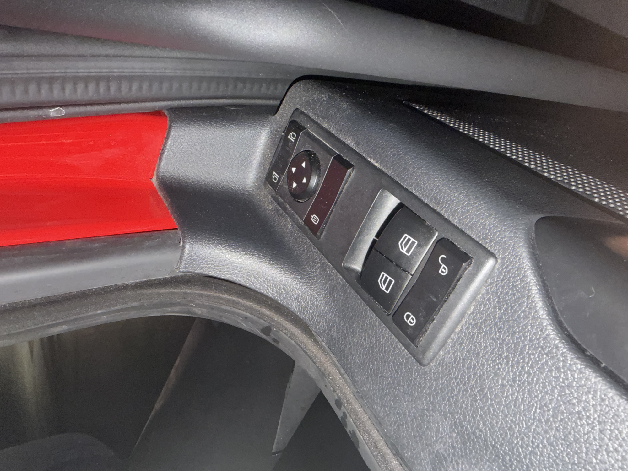 The image shows a close-up of a car's interior door panel. Dominating the frame are a set of black plastic buttons and controls. These include a directional pad with four arrows, likely for adjusting mirrors, and several other buttons with icons. To the left, there are two rectangular buttons, one with a lock icon and another with an unlock icon. Above the directional pad, there are two more rectangular buttons, one with a camera-like icon and another with a fold-out mirror icon. The car's interior appears to be black and red. The image is taken from a slightly elevated angle, looking down at the controls. There is no indication of people, actions, or interactions. The environment is limited to the car's interior. The location is stated to be the city of Wildwood, United States.