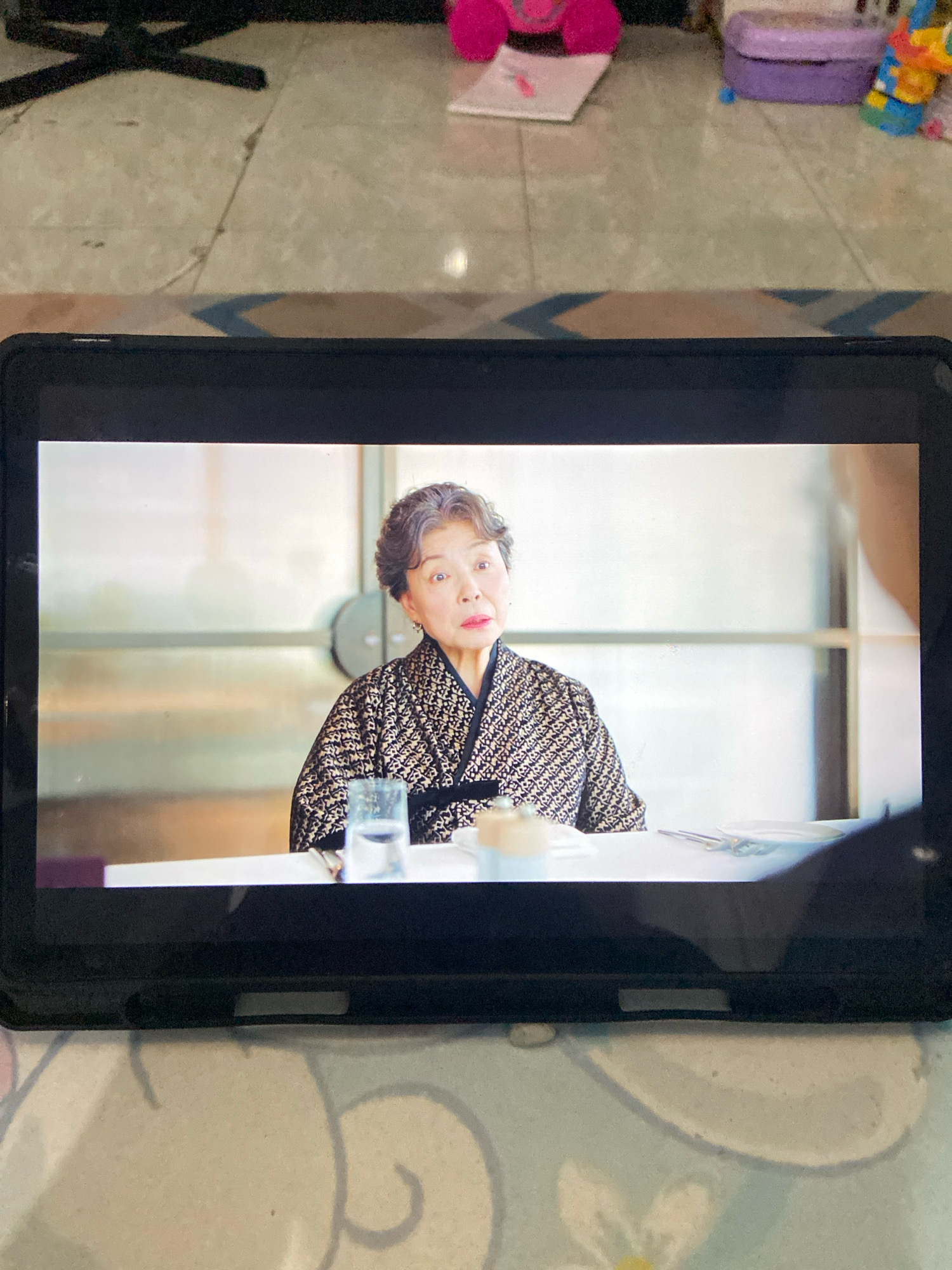 An elderly East Asian woman with gray hair and fair skin is seated at a table, looking directly at the viewer with a surprised expression. She is wearing a black and gold patterned kimono-style garment. In front of her on the table is a glass of water and a plate with cutlery. The scene is captured on a tablet screen, which is placed on a patterned surface. The background of the tablet screen shows a modern interior with a blurred background. The tablet itself is in a black case. The scene does not provide any information about the city of Gelam, Indonesia.