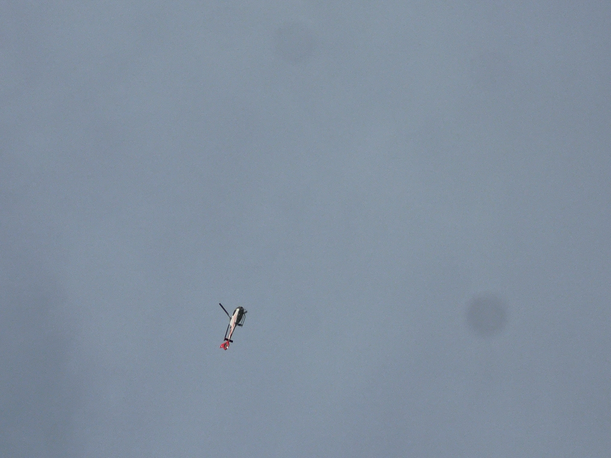 A low-angle shot reveals a lone helicopter in flight against a muted blue sky. The helicopter, positioned in the lower left quadrant of the frame, has a white body, black rotors, and red accents on its tail. A faint, blurred dark spot is visible in the sky towards the right side of the frame. The environment is nondescript, providing no clues to location within the city of Unknown, Unknown.