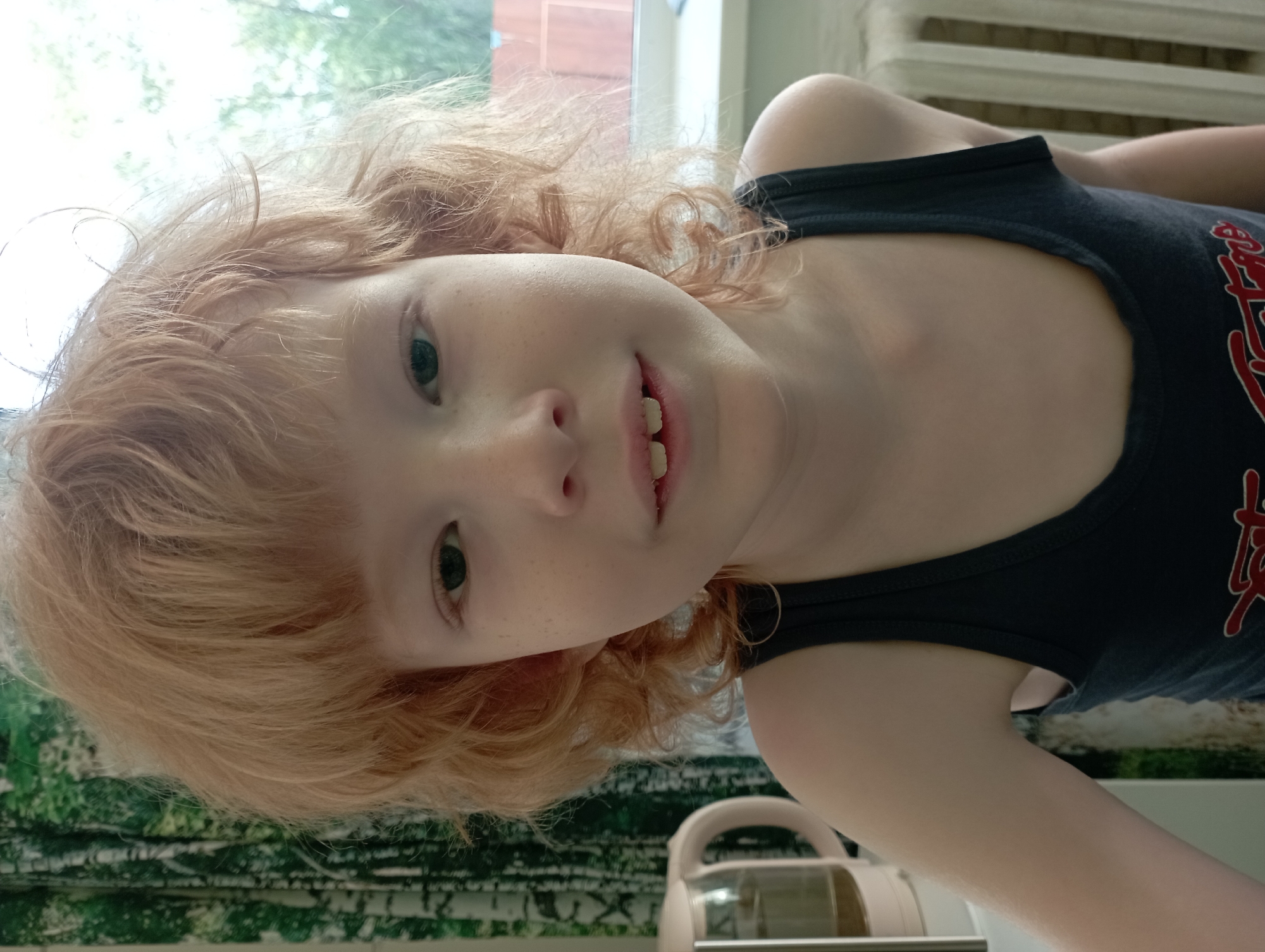 The image shows a close-up of a young child with light red, curly hair and fair skin, looking directly at the camera. The child is wearing a dark tank top with red lettering. Their face is the primary focus, with freckles visible on their cheeks. The background includes a window with a view of green trees and a glimpse of a wooden fence. To the left of the child is a white electric kettle. To the right, a radiator is visible. The scene appears to be indoors, likely in a residential setting. The image was taken in Blagoveshchensk, Russia, though no specific landmarks or context are provided to pinpoint the exact location within the city.
