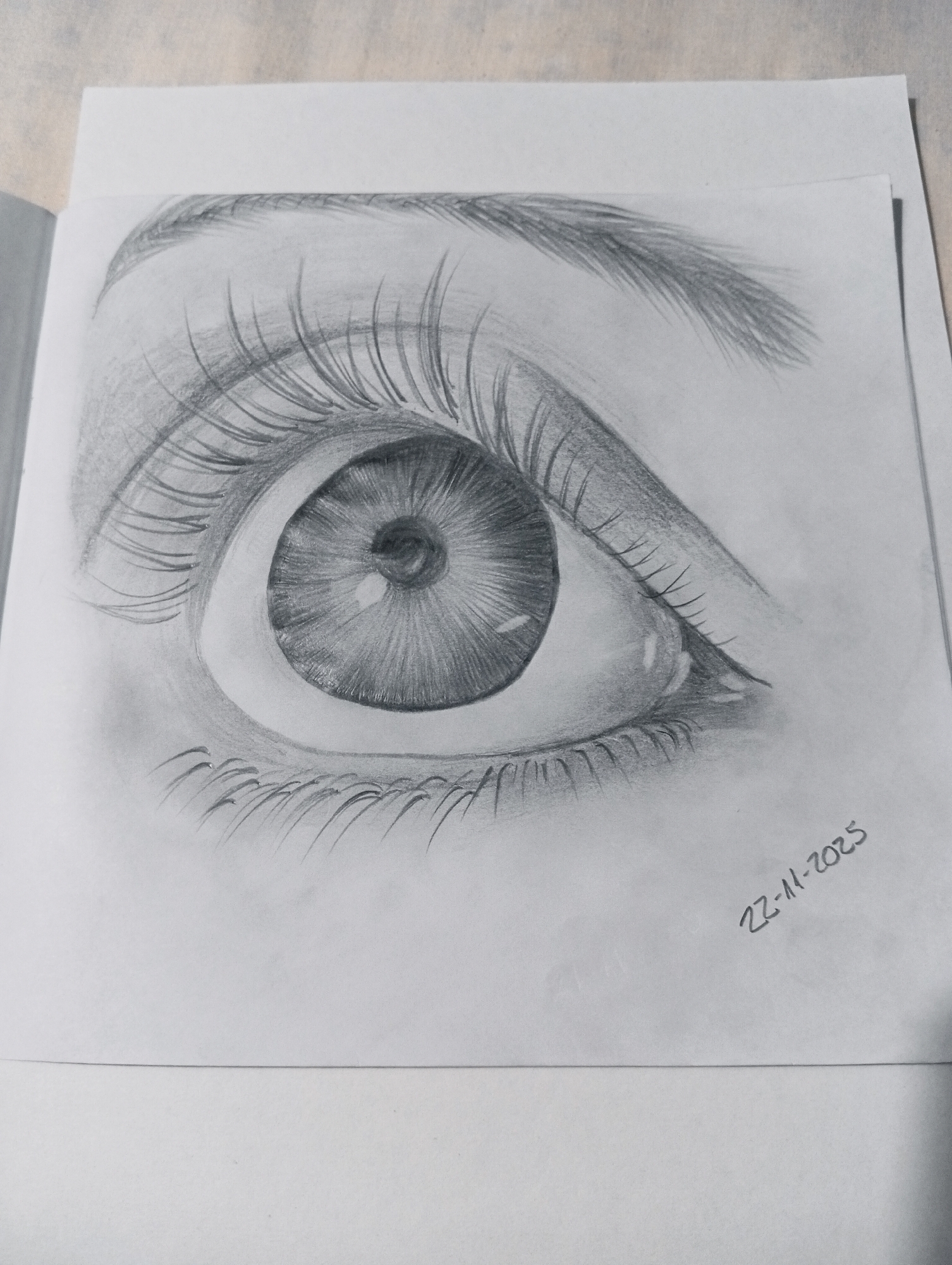 This is a black and white pencil sketch of a human eye, drawn on paper. The drawing is highly detailed, capturing the texture of the eyelashes, the iris, and the subtle shading of the eyelid. A date, "22-11-2025", is written in the lower right corner of the paper. The sketch is not located in any specific city, but rather appears to be a work of art created by an individual.