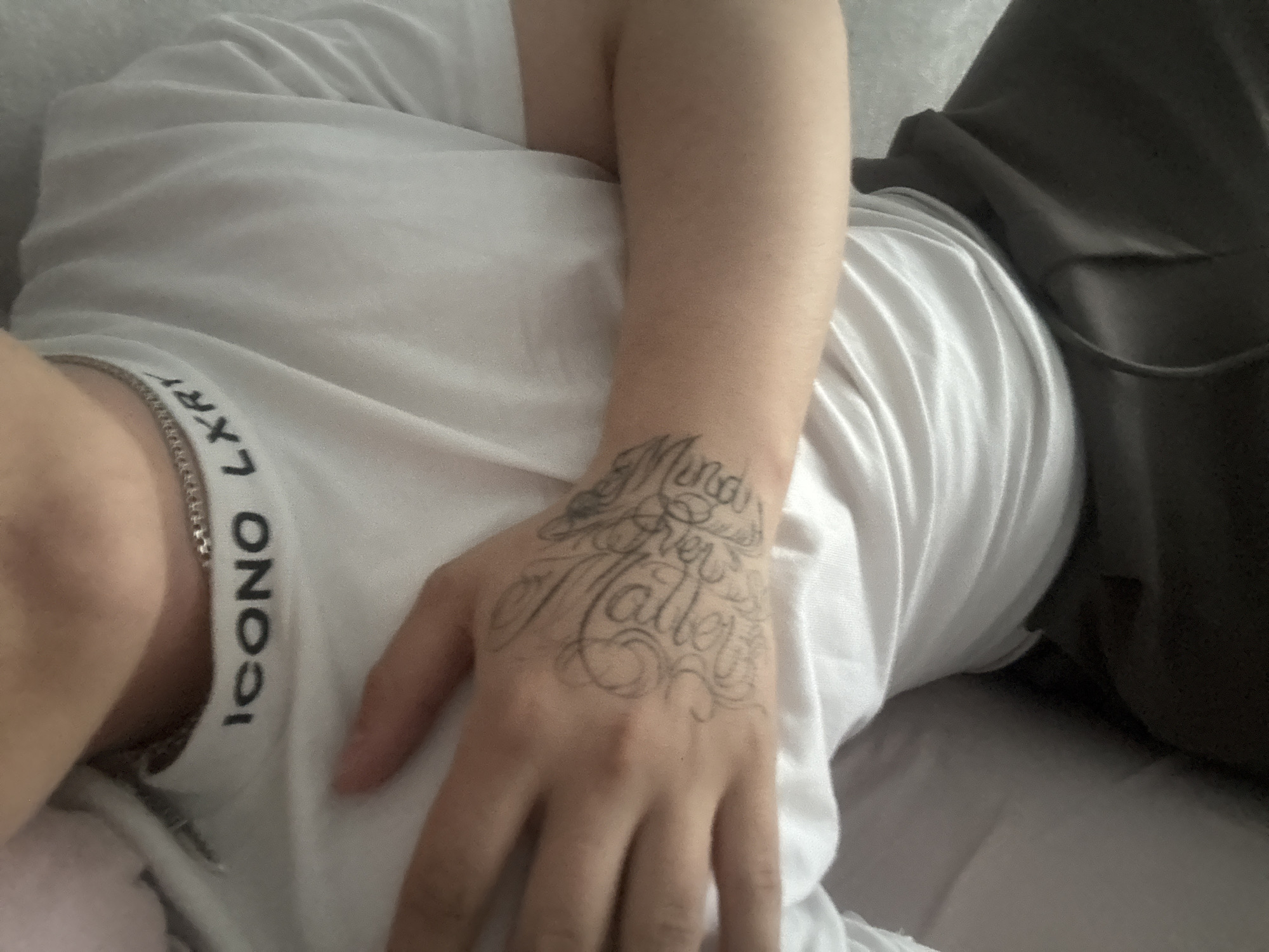 The image presents a close-up, high-angle view of a person lying down, showing their upper torso, right arm, and right hand. The individual is wearing a white t-shirt, which has visible creases, and dark-colored pants or fabric in the background. A silver chain is visible around their neck, resting against the white shirt.
The primary subject of interest is the person's right hand, which features a prominent tattoo across its back. The tattoo is rendered in a flowing, calligraphic script and clearly reads "Mind Over Matter," with decorative swirls and flourishes surrounding the text.
On the collar area of the white t-shirt, some text is visible. The word "ICONO" can be clearly read, alongside partially visible letters that appear to be "LXR" or "LXRT."
The setting appears to be indoors, suggested by the soft, diffused lighting and the relaxed posture of the person, possibly on a bed or couch. No specific activity is taking place beyond what appears to be resting or relaxing. The time of day and exact location are not discernible from the image.