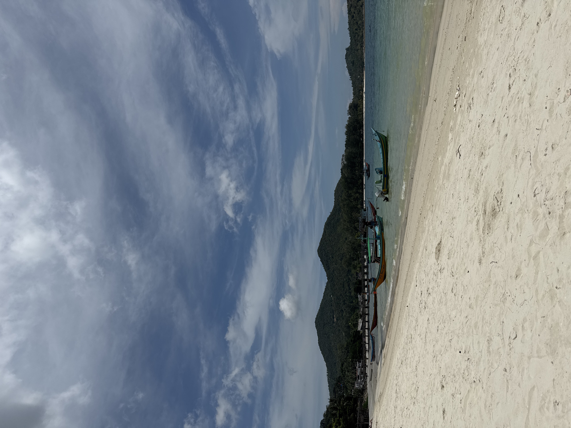 The image shows a beach in Baan Chaloklum, Thailand. The beach has light sand, and the water is clear and shallow. A number of traditional Thai longtail boats are moored along the shoreline. The background includes lush, green hills, suggesting a tropical environment. The sky is cloudy. There are no visible people in the immediate scene.