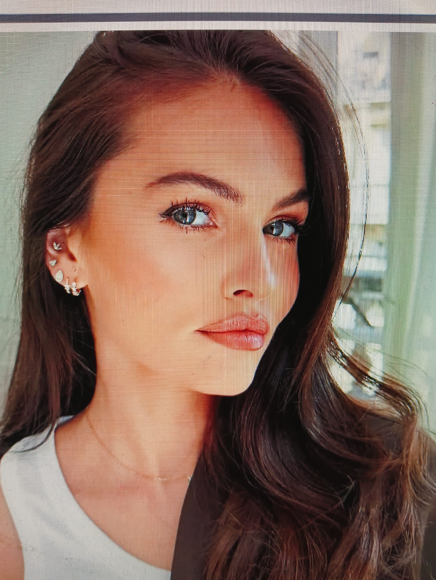 The image shows a close-up portrait of a woman with long, wavy brown hair. She is wearing a white top and a dark jacket. Her left ear is adorned with multiple earrings, including small studs and a larger anchor-shaped earring. She is also wearing a delicate gold necklace. The background appears to be an indoor setting with soft lighting and out-of-focus elements suggesting a room. There are no discernible landmarks or details within the image that would allow for the identification of a specific location in Sluck, Belarus.
