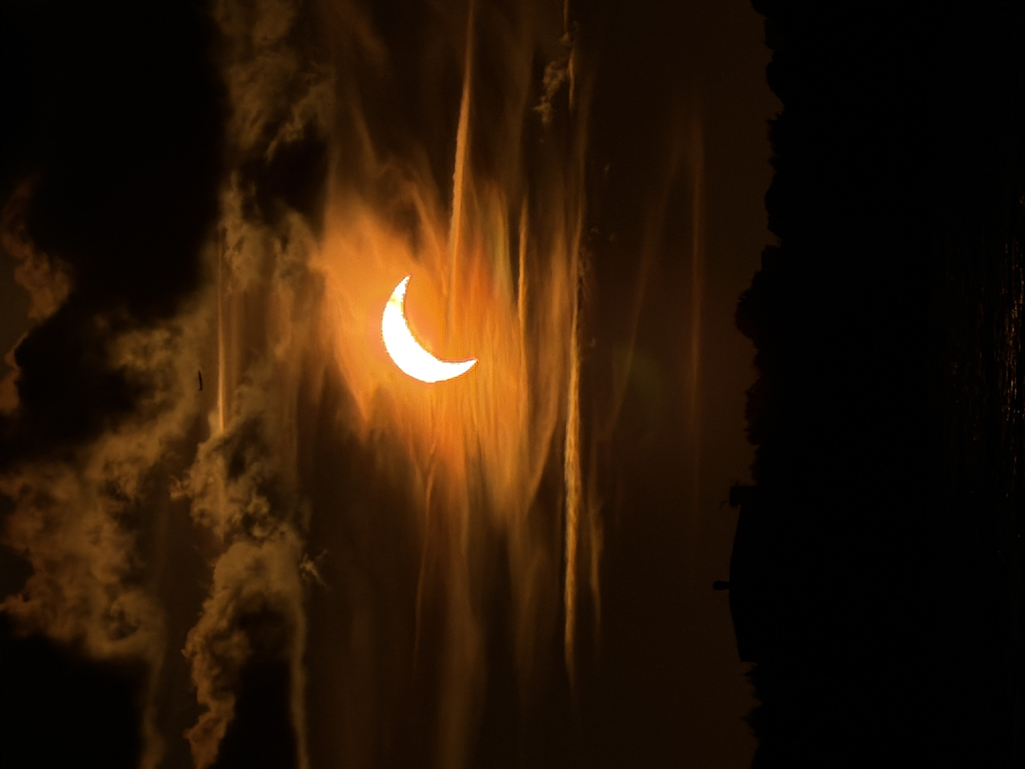 This is a vertical, telephoto photograph of a solar eclipse observed from Riga, Latvia. The sun, partially obscured by the moon, appears as a brilliant crescent against a backdrop of dramatic, textured clouds illuminated with a warm, orange glow. A small airplane is visible in the upper portion of the sky. The foreground is dominated by a silhouetted landscape, featuring the dark outline of trees and a building, possibly a house with a chimney. The lower part of the image shows the dark, reflective surface of water. The overall impression is one of a celestial event occurring during sunset or sunrise.