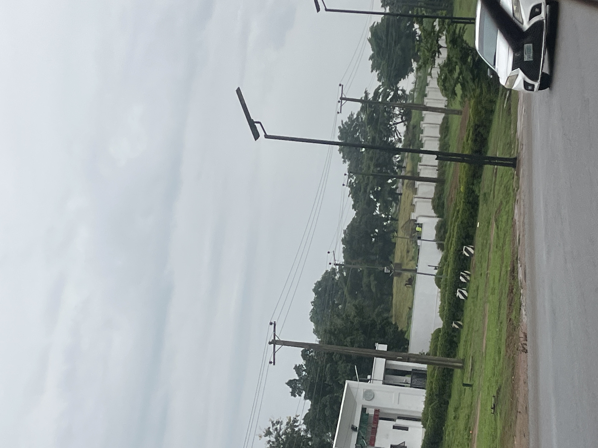 The media file captures a street scene in Kuchingoro, Nigeria, under an overcast sky, suggesting a cloudy day or early morning/late afternoon light. The primary subjects are several tall, dark streetlights positioned along the roadside, interspersed with utility poles carrying power lines. A white car is partially visible on the right side of the frame, parked on a wet asphalt road, indicating recent rain or damp conditions.
Beyond the road and streetlights, a white building with a modern architectural style is partially visible. Adjacent to the building and stretching into the background is a lush green expanse of grass and trees, with a white wall or fence line visible intermittently. Several striped, possibly decorative, objects are scattered across the grassy area.
The scene appears to be a quiet, possibly residential or commercial area, given the presence of the building and streetlights. No specific activity or event is clearly depicted, but the wet road and overcast sky create a somewhat subdued atmosphere. There is no visible text within the image.